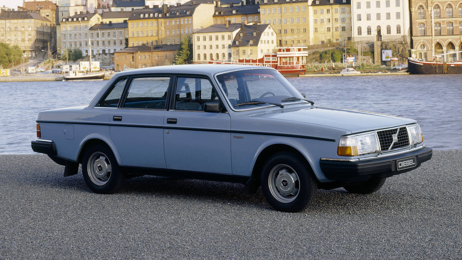 Volvo iPhone城市河畔風景 壁紙 — 一輛淺藍色富豪240柴油房車停泊在海濱的碎石路上，背景有歷史建築和船隻。