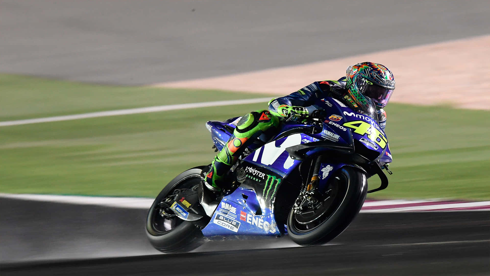 VR46 2018雅馬哈原型機車 壁紙 — 一名電單車賽車手在人工燈光下的賽道上，身穿鮮豔的藍、綠、紫色裝備，傾斜入彎。