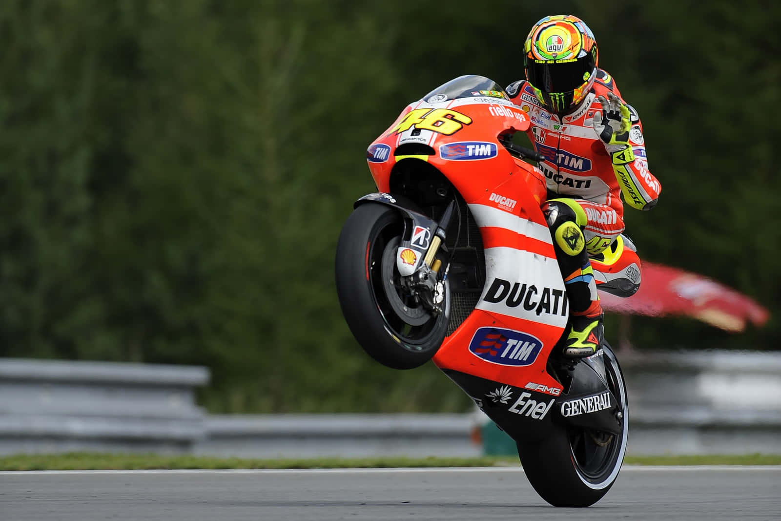 VR46翹起杜卡迪機車 壁紙 — 一位身穿紅白杜卡迪賽車服的電單車車手在賽道上表演前輪離地，背景有綠樹。