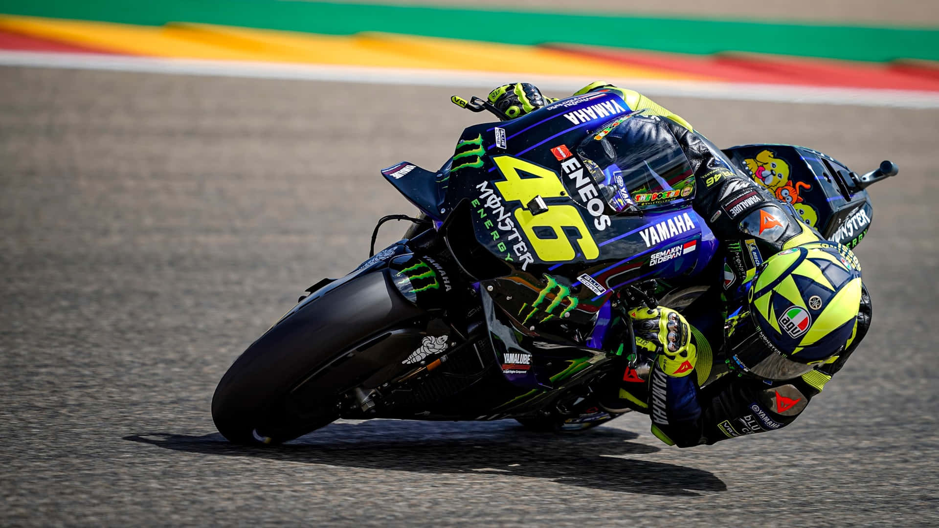 VR46 意大利職業MotoGP賽車手 壁紙 — 一名電單車賽車手在陽光明媚的賽道上急彎傾側，身穿鮮豔的藍黃色賽車服。