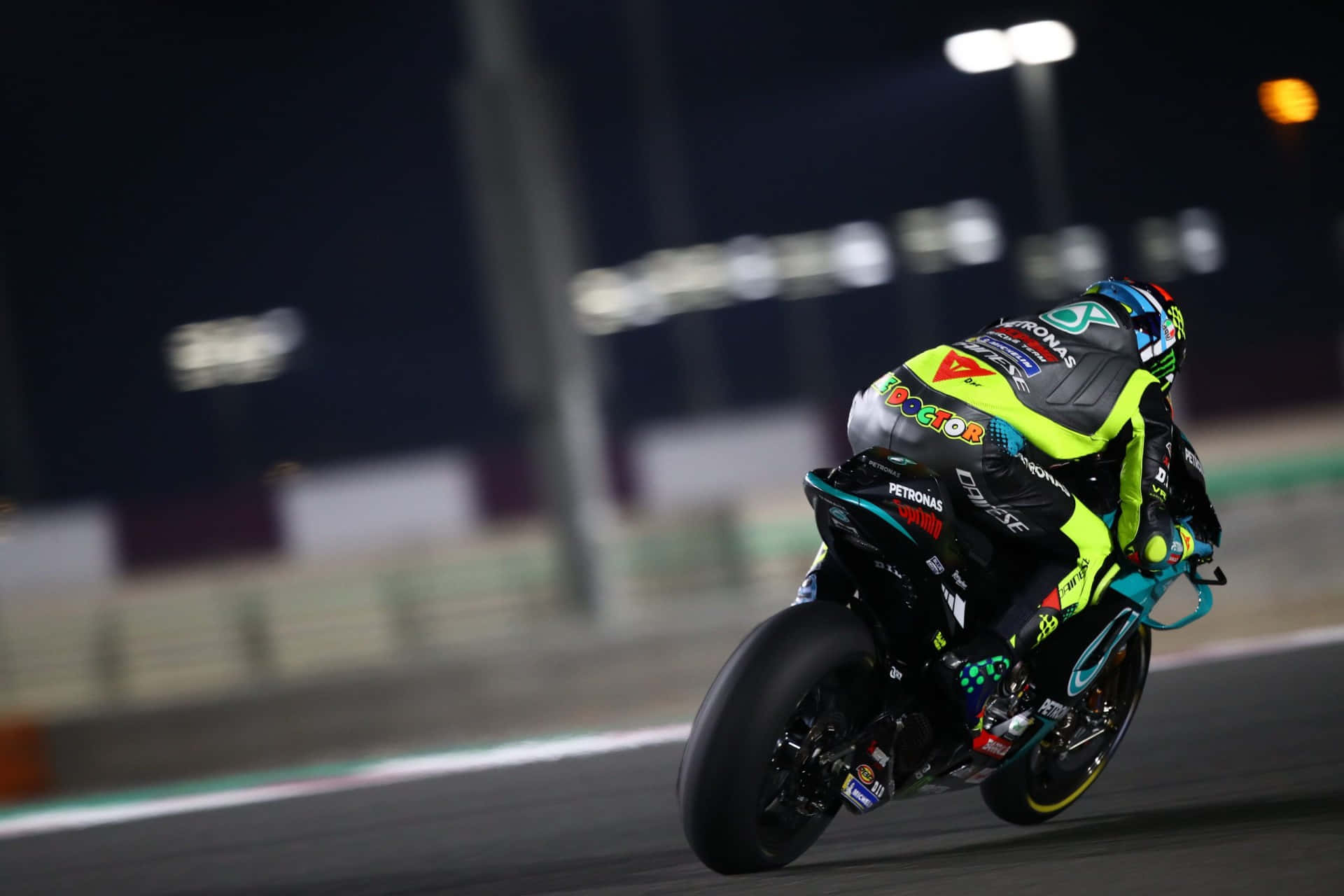 VR46 與Petronas Yamaha賽車隊聯名賽事 壁紙 — 一名電單車手在夜間燈火通明的賽道上傾斜入一個急彎，背景是模糊的體育場燈光。