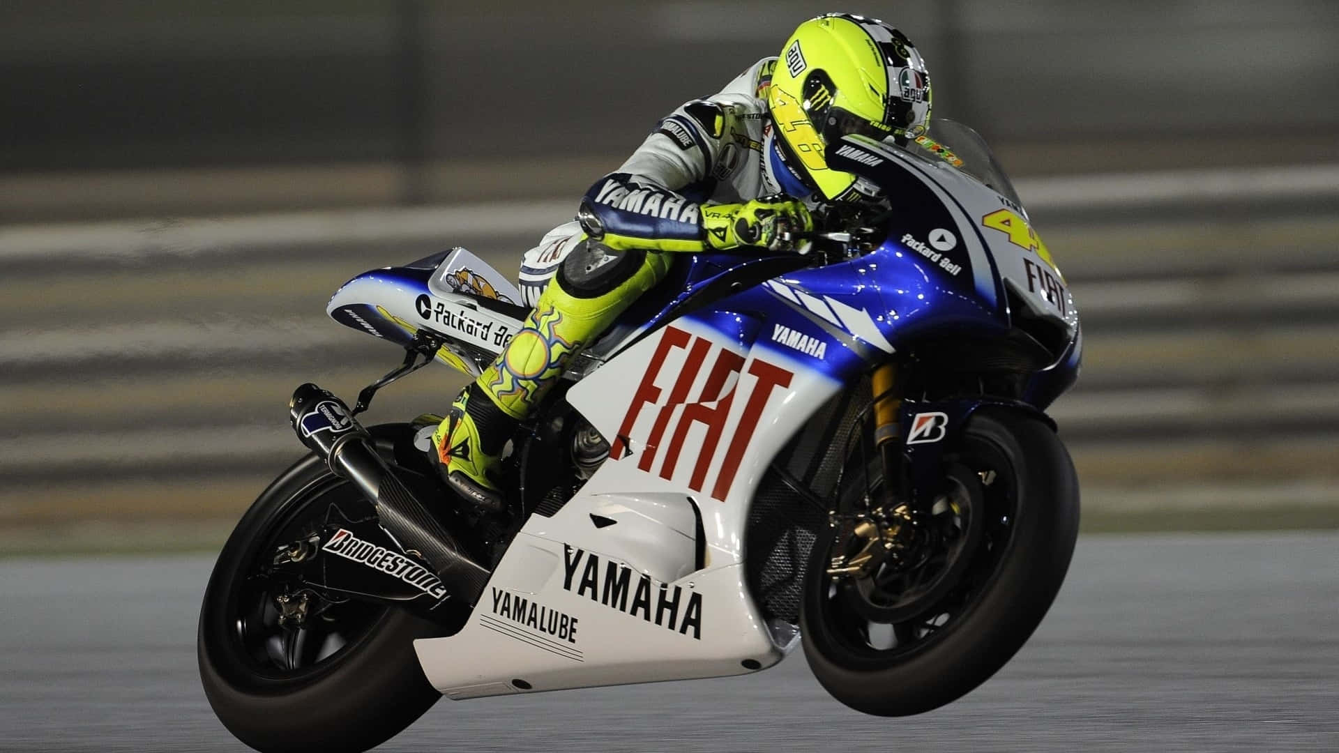 VR46 菲亞特雅馬哈超級賽車 壁紙 — 一名電單車手在賽道燈光下，騎著Yamaha賽車傾斜入彎，背景呈現動態模糊效果。