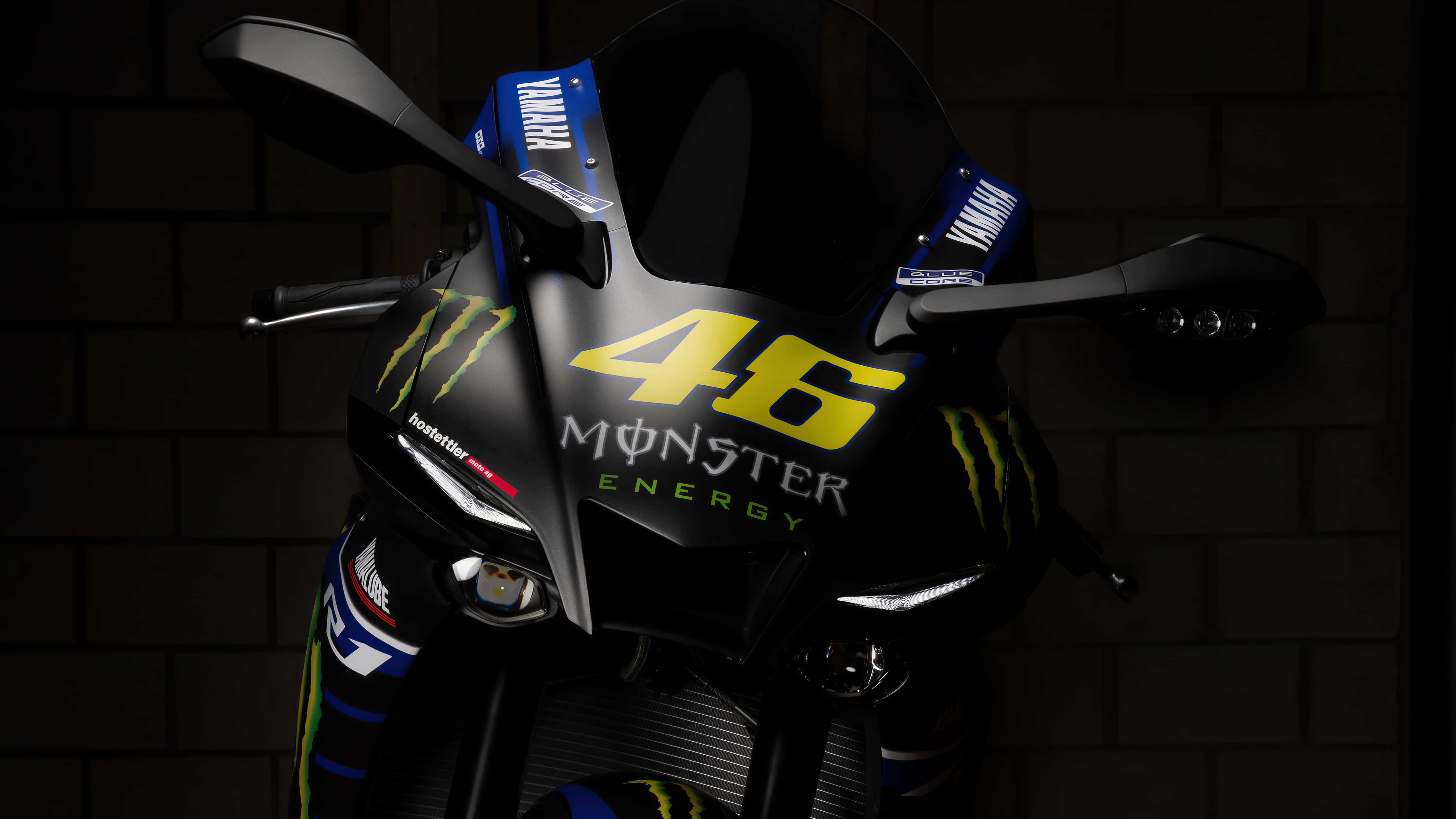 VR46 雅馬哈賽車配備 Monster Energy 飲料標誌 壁紙 — 一架型格黑色Yamaha電單車，帶有搶眼黃色46 Monster Energy品牌標誌，在深色磚牆前被戲劇性地照亮。