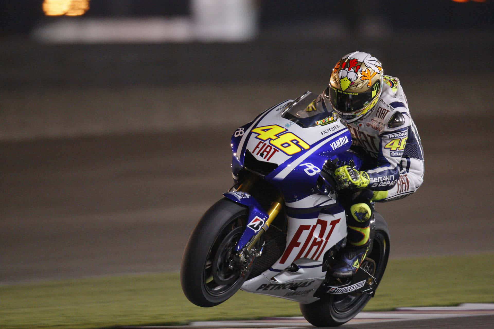 VR46 騎乘菲亞特賽車於卡塔爾MotoGP賽前測試 壁紙 — 一位電單車手在夜間燈火通明的賽道上傾斜入彎，戴著色彩繽紛的頭盔和賽車服。