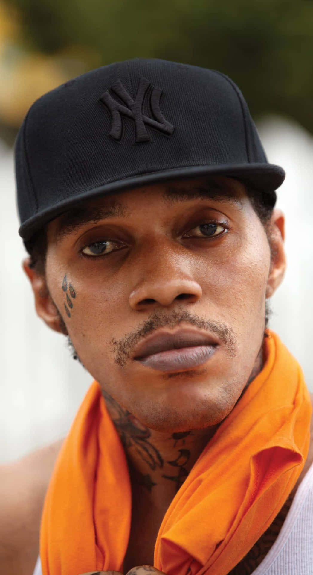 Vybz Kartel 經典造型 壁紙 — 一個紋身男子的特寫肖像，戴著黑色NY帽子和鮮豔的橙色圍巾。