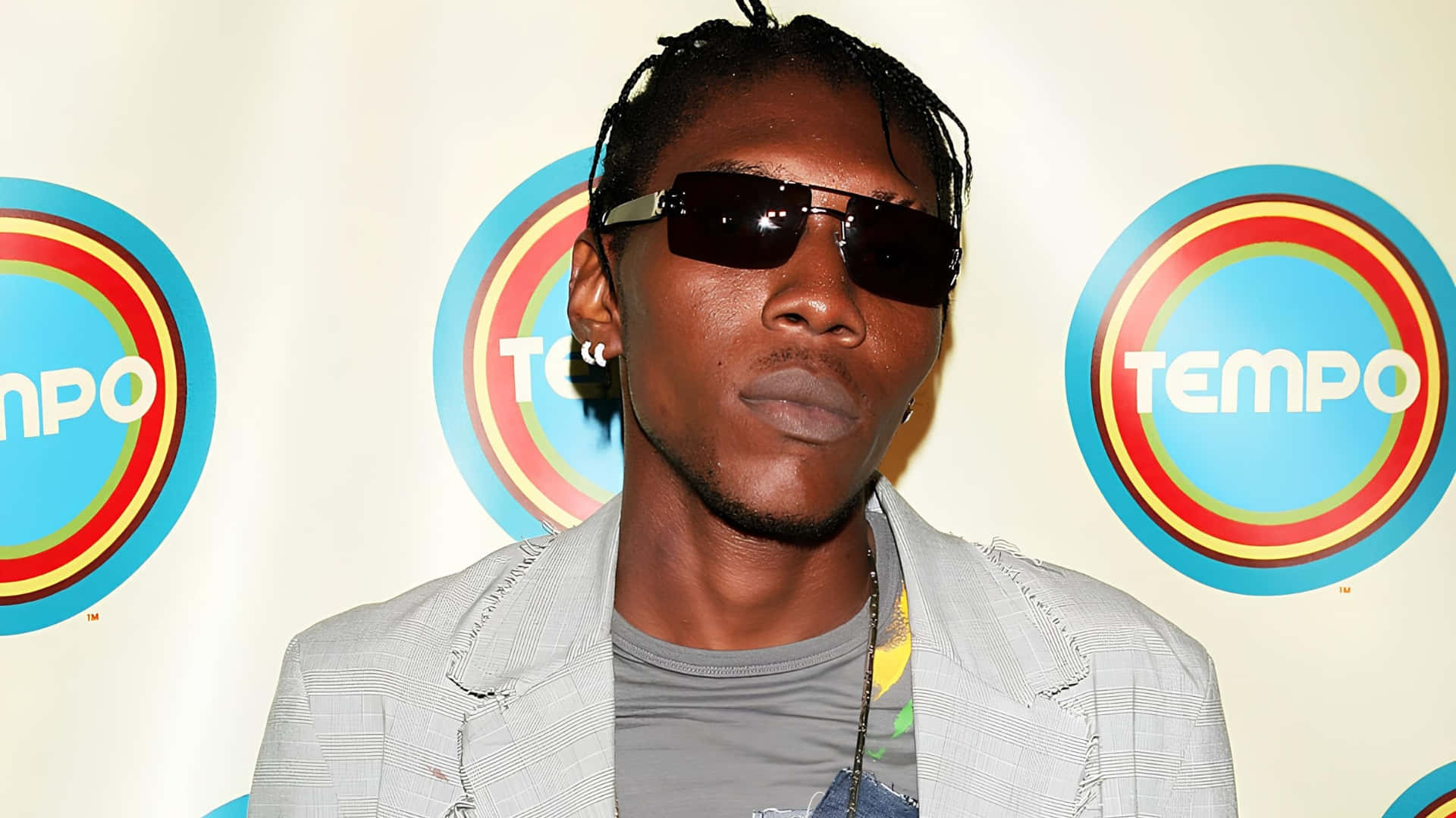 Vybz Kartel 風格偶像 壁紙 — 一名辮子頭髮、戴著深色太陽眼鏡的男子，在色彩繽紛的Tempo標誌背景前自信地擺姿勢。