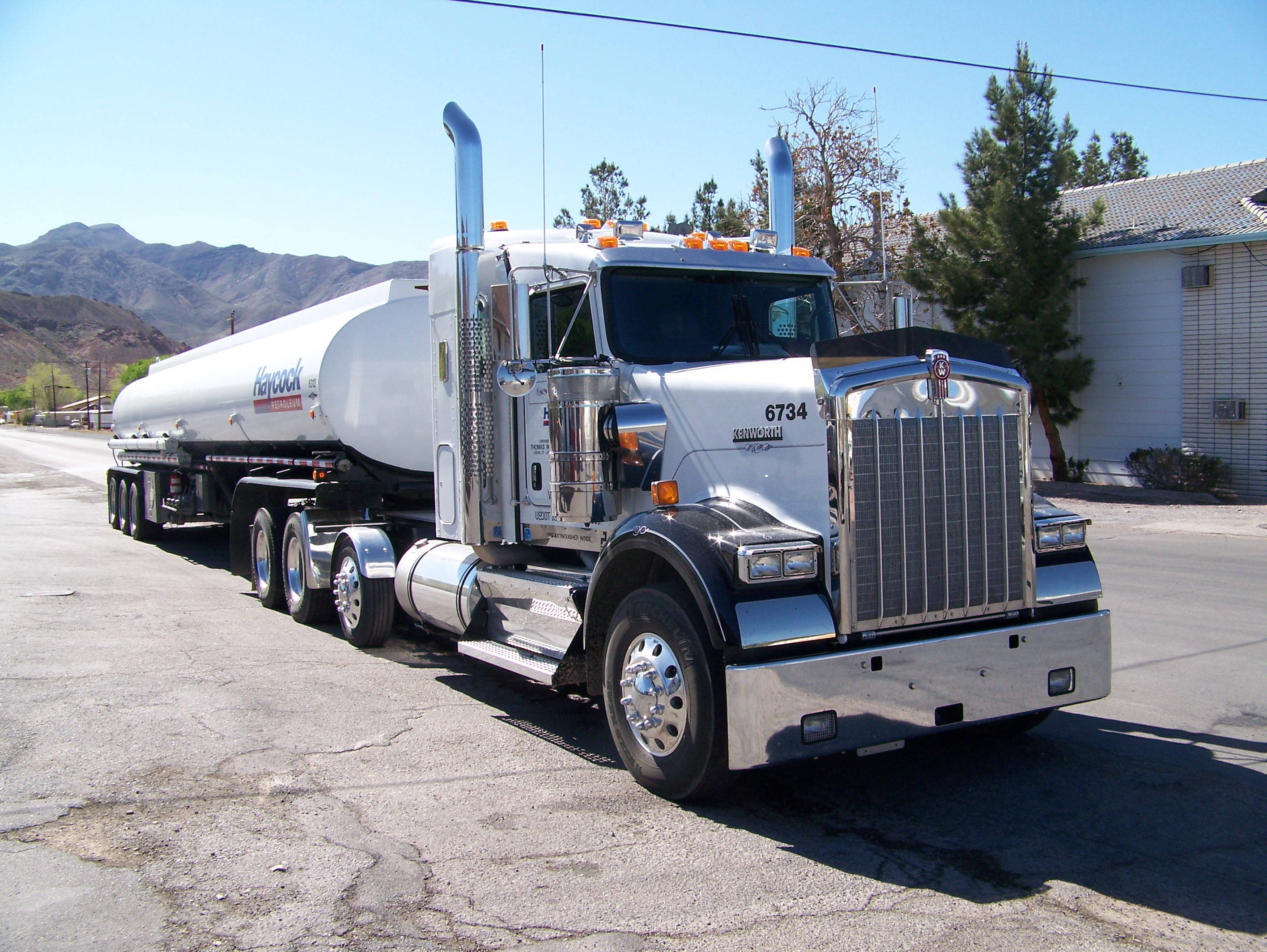 肯沃斯 W900 油罐車 壁紙 — 一輛閃亮的白色Kenworth油罐車，帶有鍍鉻裝飾，停在裂開的瀝青路上，背景是晴朗的藍天和山脈。
