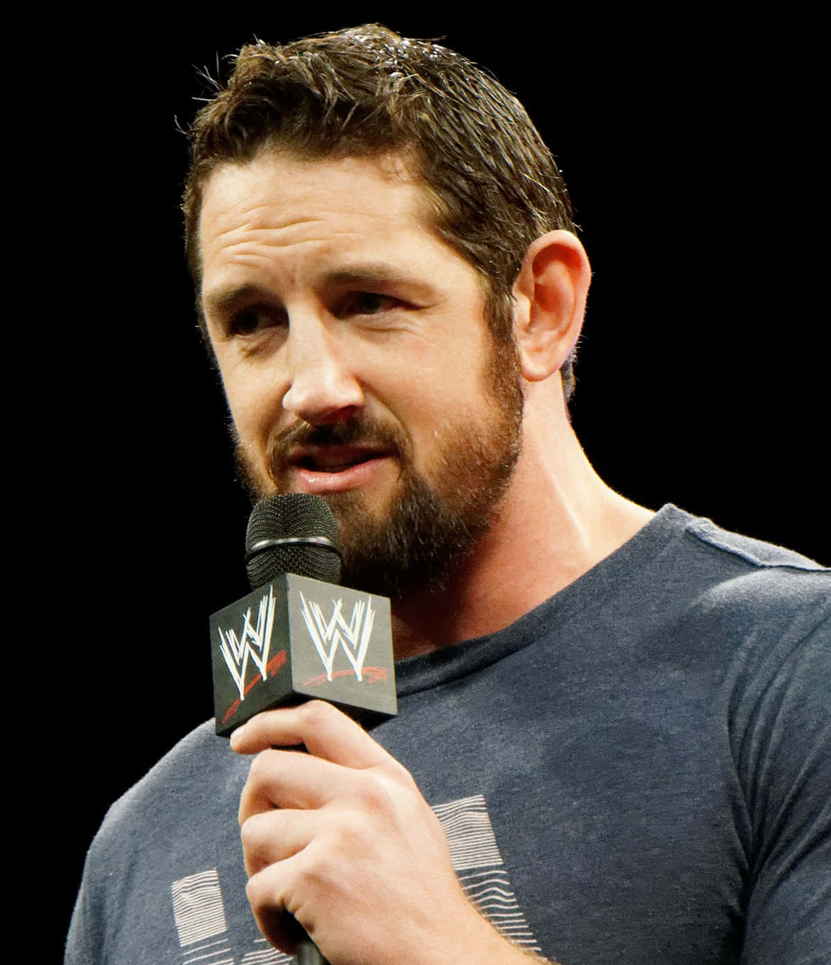 Wade Barrett 拿著麥克風 壁紙 — 一個留鬍鬚的男人穿著深色T恤，在黑色背景前拿著WWE麥克風。