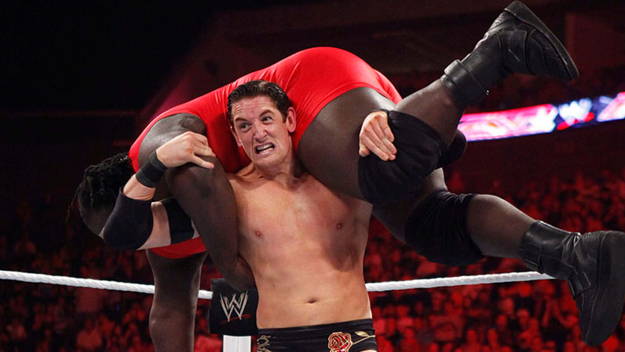 Wade Barrett背負Mark Henry 壁紙 — 身穿黑色短褲的摔角手在明亮燈光下的摔角場中，將身穿紅色緊身衣的對手舉過肩膀。