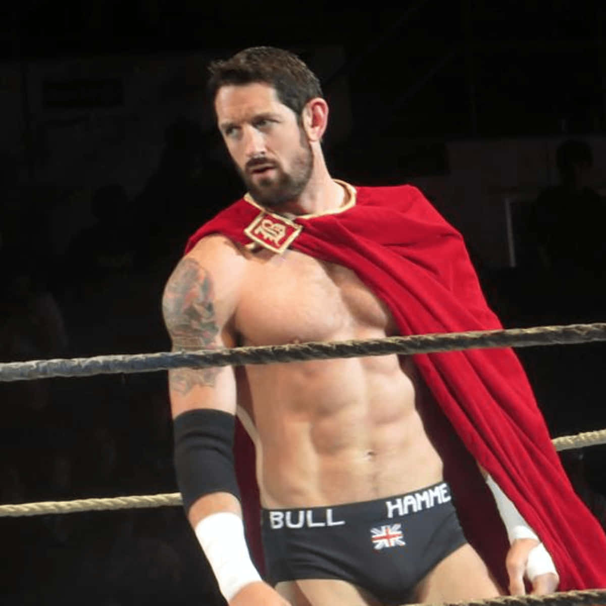 Wade Barrett身穿披風 壁紙 — 一位肌肉發達的摔角手，披著紅色斗篷，手臂上有紋身，站在燈光璀璨的摔角擂台上。
