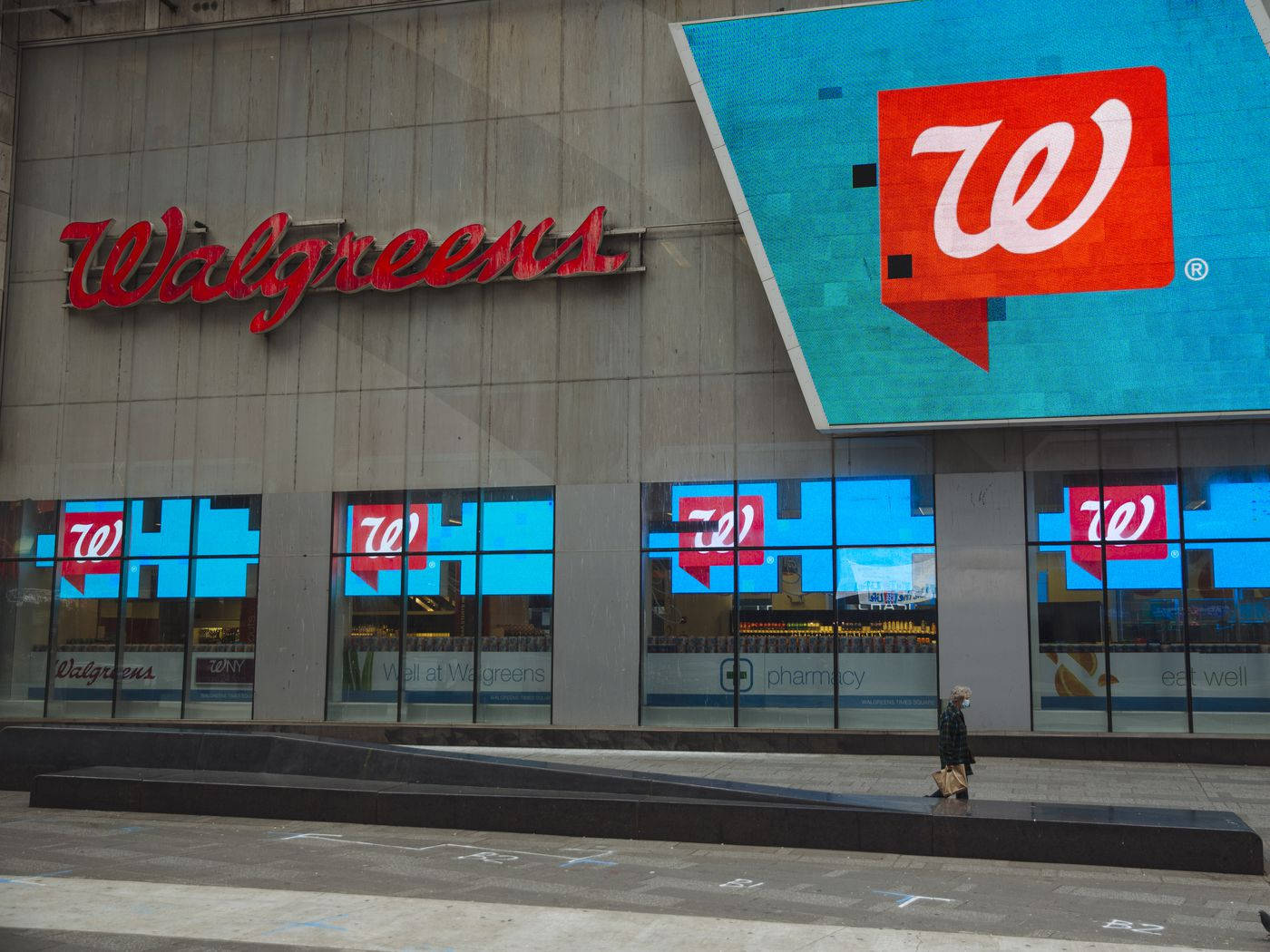 Walgreens藥房門店正面建築 壁紙 — 一個人走過一間Walgreens商店，商店有醒目的紅色招牌和反映城市環境的藍色玻璃窗。