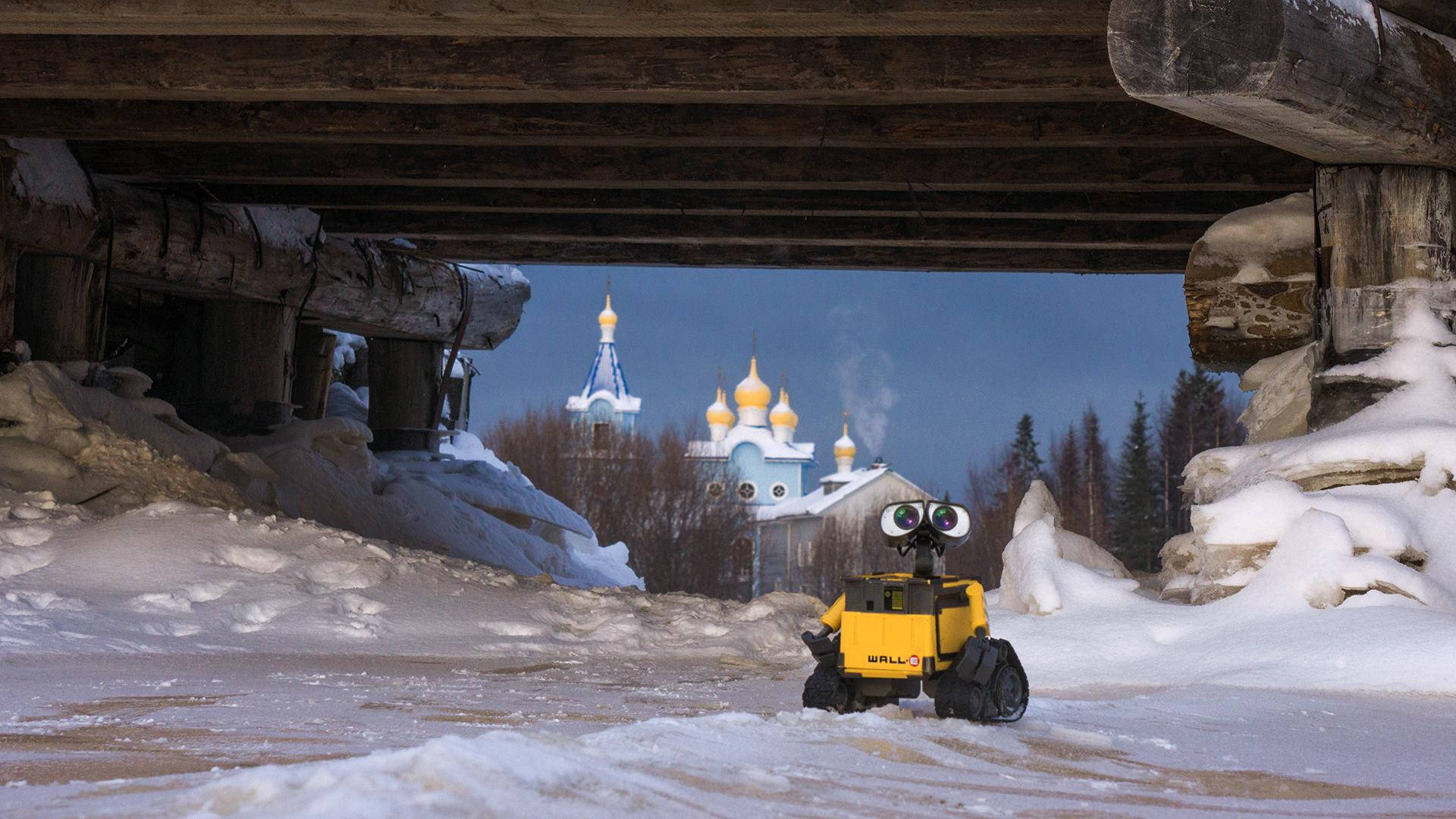 橋下 WALL·E 壁紙 — 一個黃色機械人，有大眼睛，企喺落雪嘅地下通道，望住遠處洋蔥頂教堂，背景係黃昏天空。