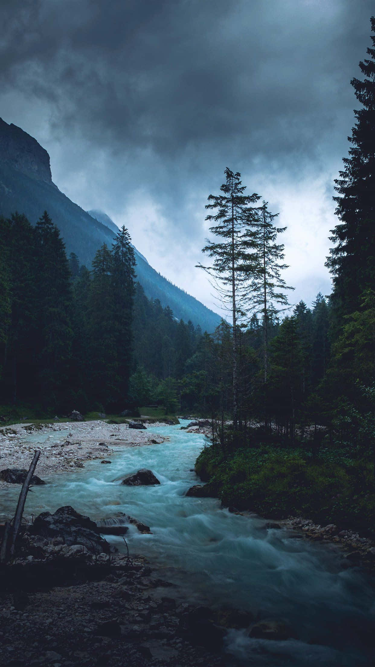 Dark Nature 桌布 壁紙 — 一條湍急的河流在暴風雨灰暗的天空下，穿過陰暗的森林山谷，背景是高聳的山脈。