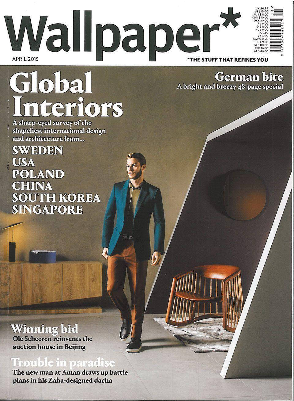 Wallpaper Global Interiors 雜誌封面 壁紙 — 一位穿著青綠色西裝外套的時尚男士走過一個配有木製家具和幾何牆壁的現代室內空間。