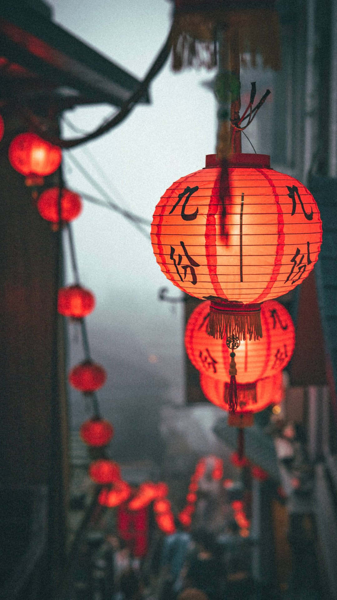  壁紙 — 溫暖發光的紅色紙燈籠，帶有黑色書法，懸掛在黃昏時分霧氣瀰漫的小巷中。
