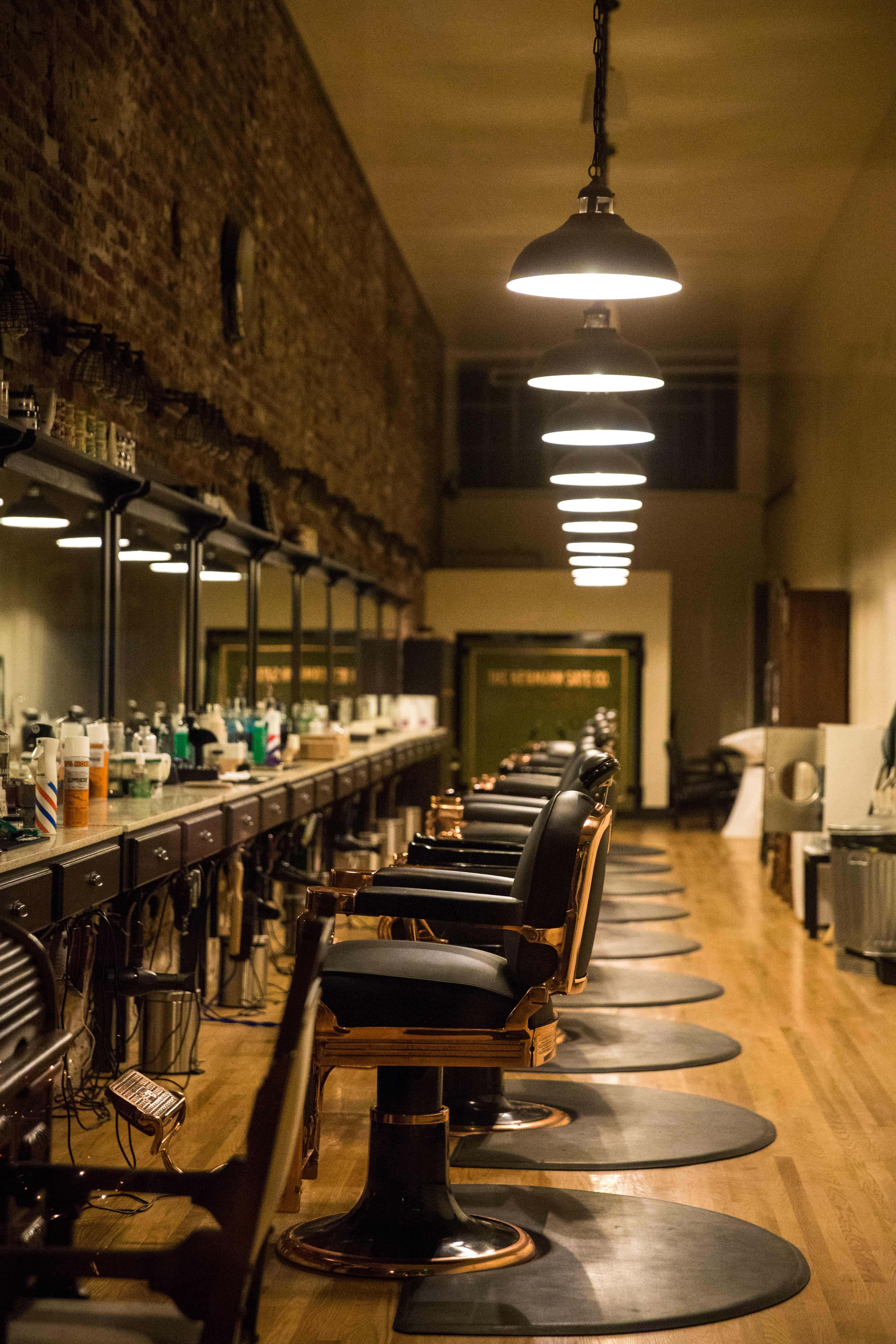 Barber 桌布 壁紙 — 一間燈光溫暖的復古理髮店，舖有木地板、黑色理髮椅，以及掛滿鏡子和護理產品的裸露磚牆。