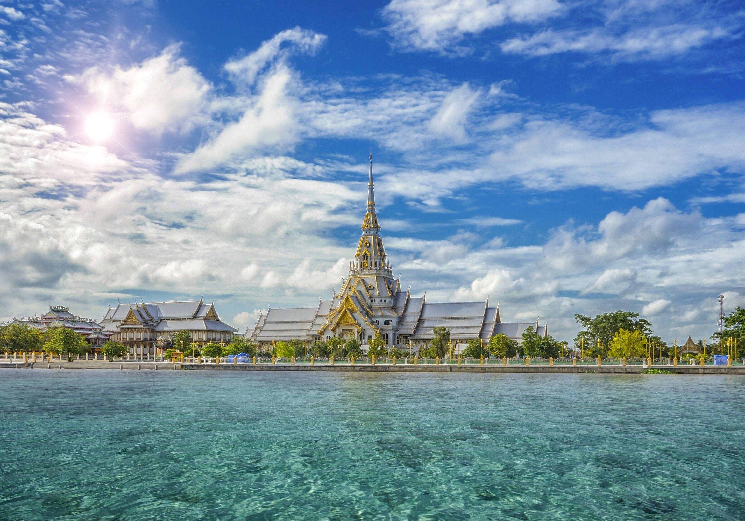 泰國 Wat Sothonwararam 寺廟 壁紙 — 一座宏偉的泰國寺廟，擁有金色尖頂和白色屋頂，雄偉地矗立在清澈的藍綠色水邊，上方是明亮的藍天和蓬鬆的白雲。