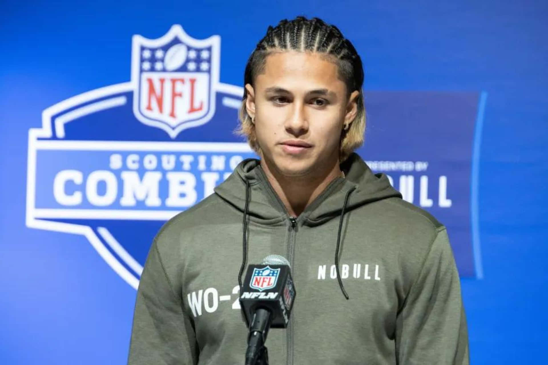  壁紙 — 一位辮子頭髮的年輕NFL球員在Scouting Combine記者會上穿著灰色連帽衫站在麥克風前。