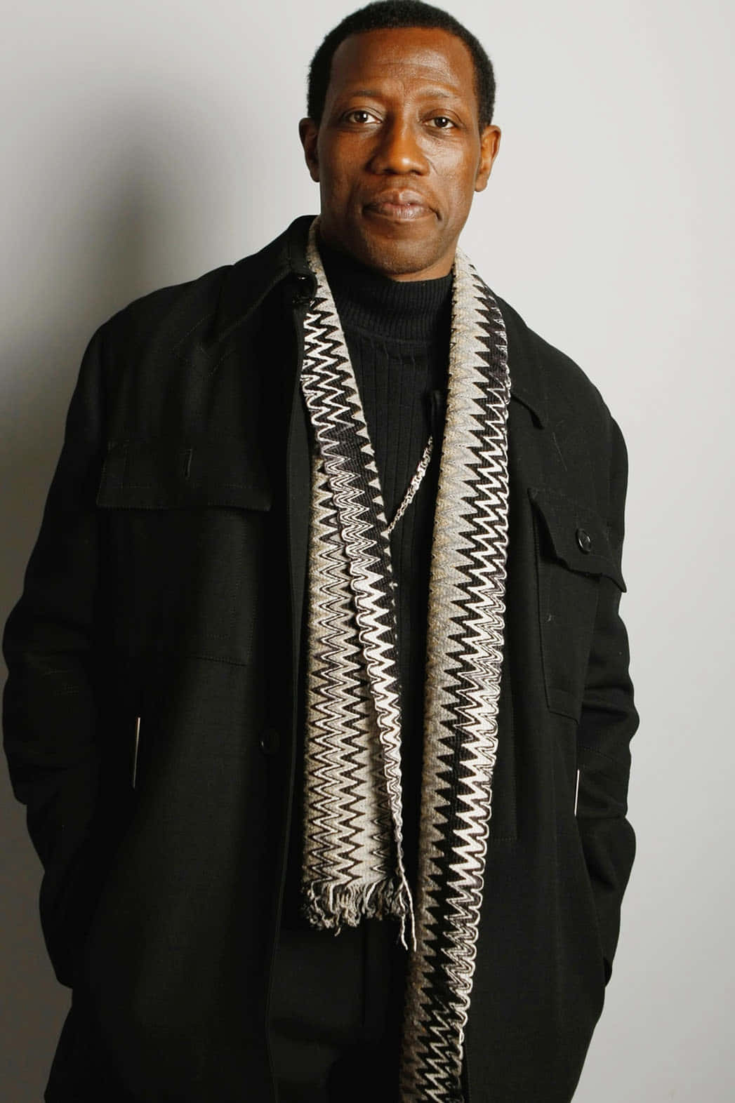 Wesley Snipes 桌布 壁紙 — 一位穿著黑色外套和圖案圍巾的沉穩男子，雙手插袋，站在素淨的灰色牆前。