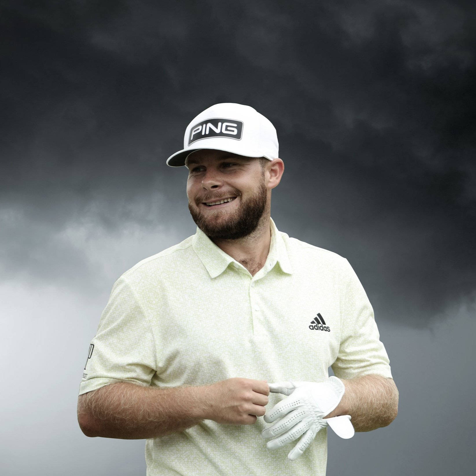 Tyrrell Hatton 桌布 壁紙 — 一位戴白帽穿淺色襯衫微笑的高爾夫球手，在戲劇性的暴風雨天空下拿著手套。