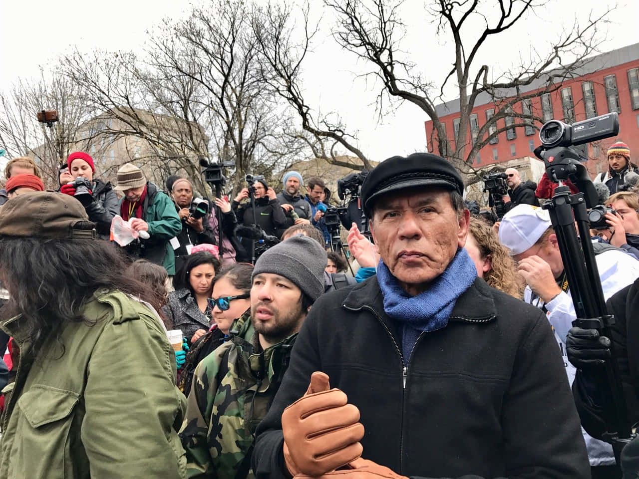  壁紙 — 一位戴黑色鴨舌帽和藍色圍巾的年長男子在公眾抗議活動中堅定地站著，被記者和攝影機包圍。