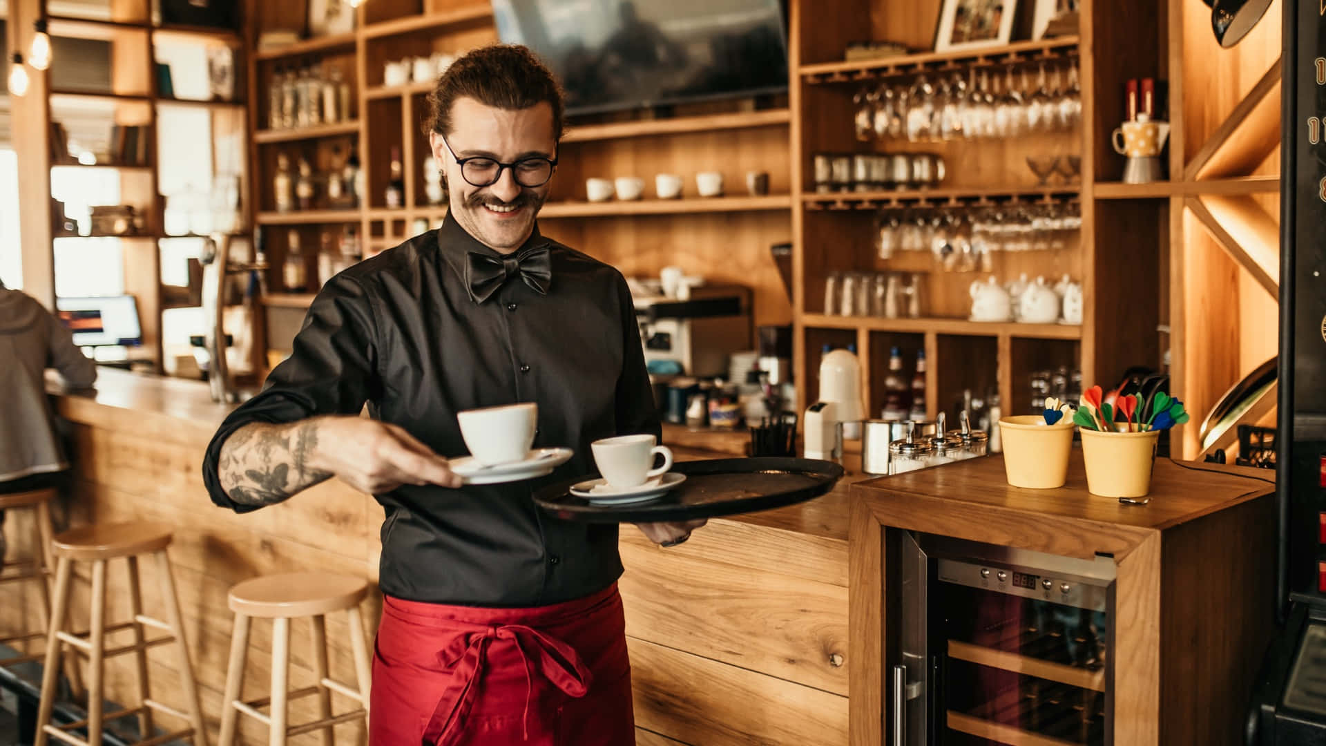 Coffee Shop 桌布 壁紙 — 一位戴眼鏡、身穿紅色圍裙的微笑咖啡師，在溫暖燈光的木質咖啡館中，用托盤端著兩杯咖啡。