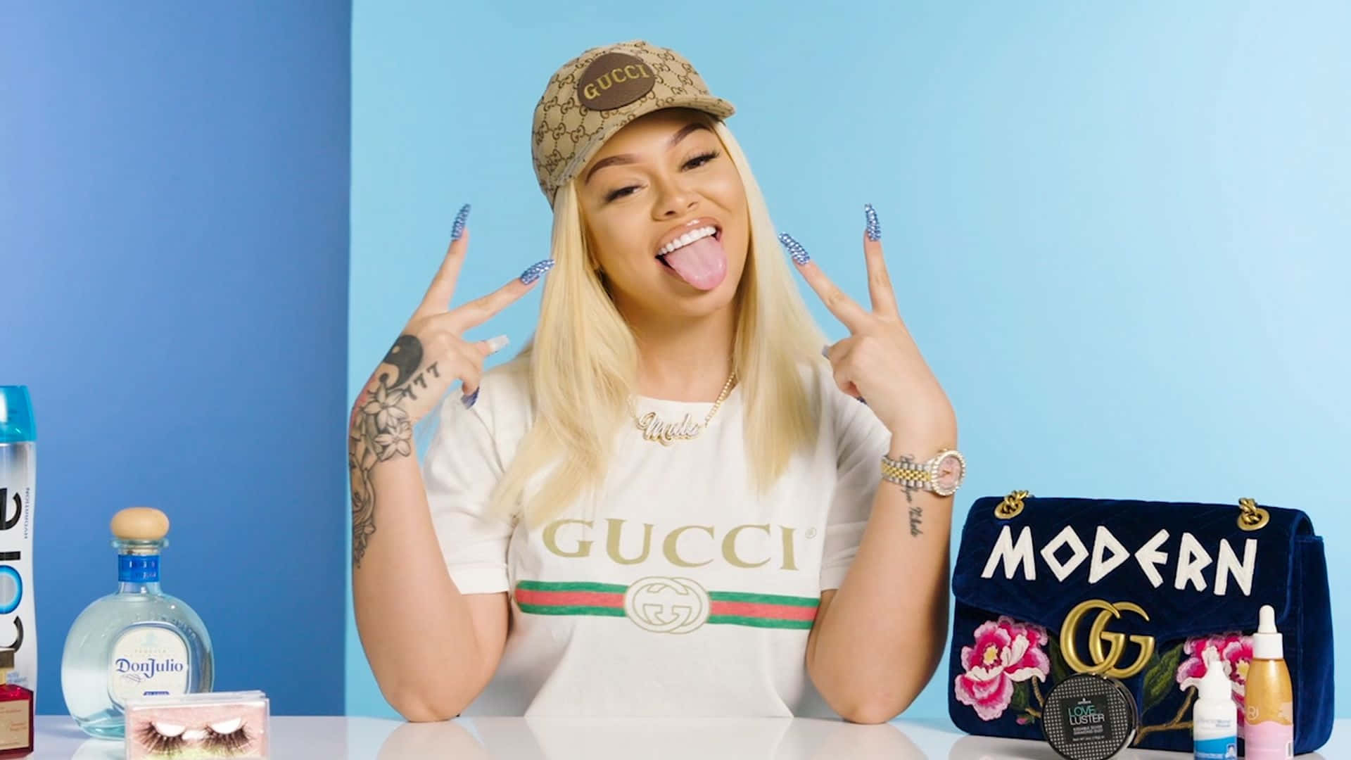  壁紙 — 一位金髮女子戴著Gucci帽子和穿著Gucci襯衫，頑皮地伸出舌頭，同時用閃亮指甲做出和平手勢。