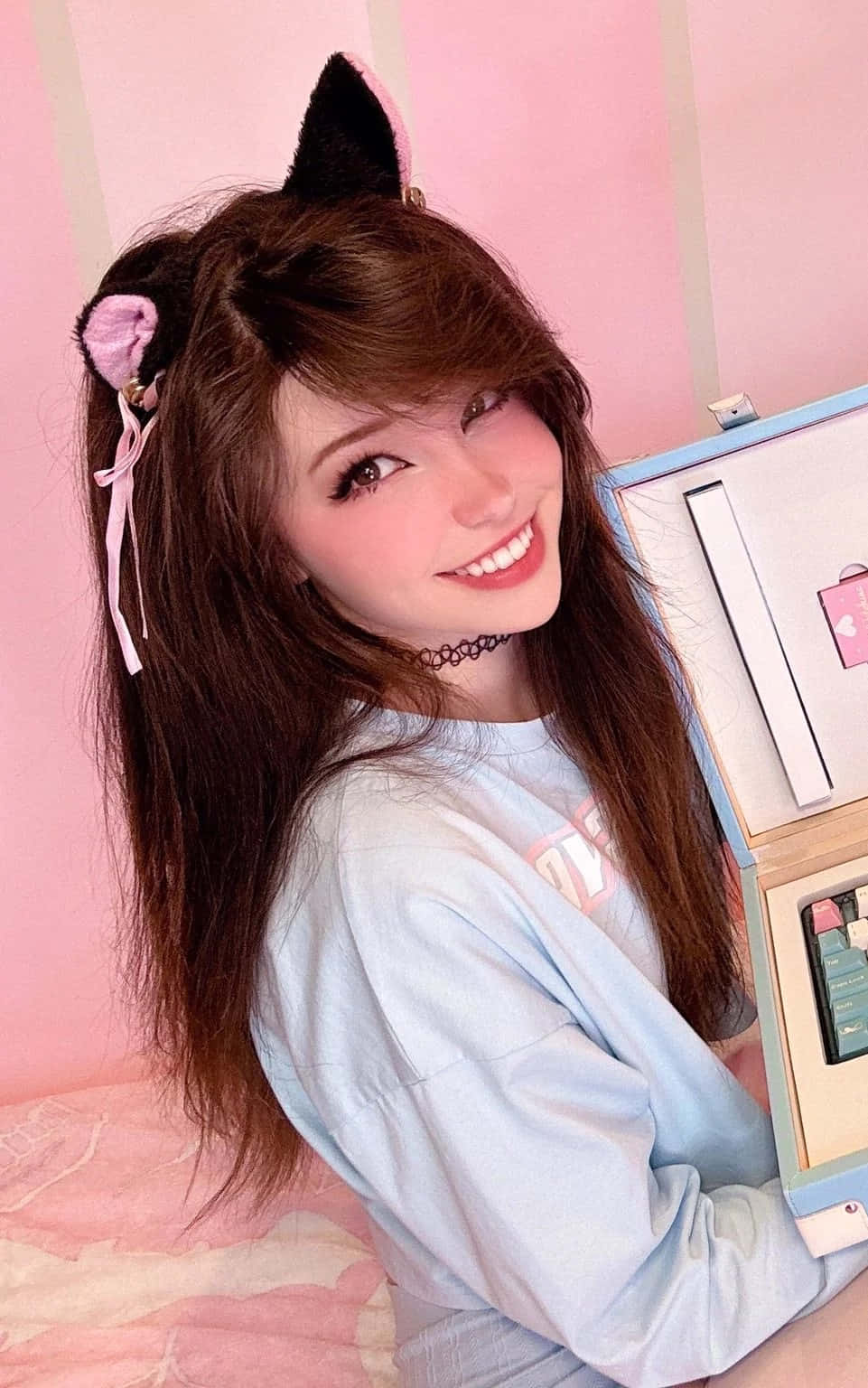  壁紙 — 一位面帶微笑、戴著貓耳朵、留著棕色長髮的年輕女子，在粉紅色條紋牆前開心地擺姿勢。