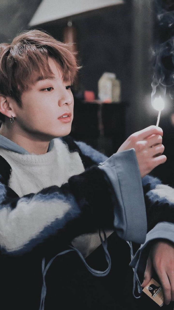 BTS 田柾國 可愛火柴 壁紙 — 一位淺棕色頭髮的年輕男子手持一根點燃的火柴，在黑暗背景下散發出溫暖的光芒。