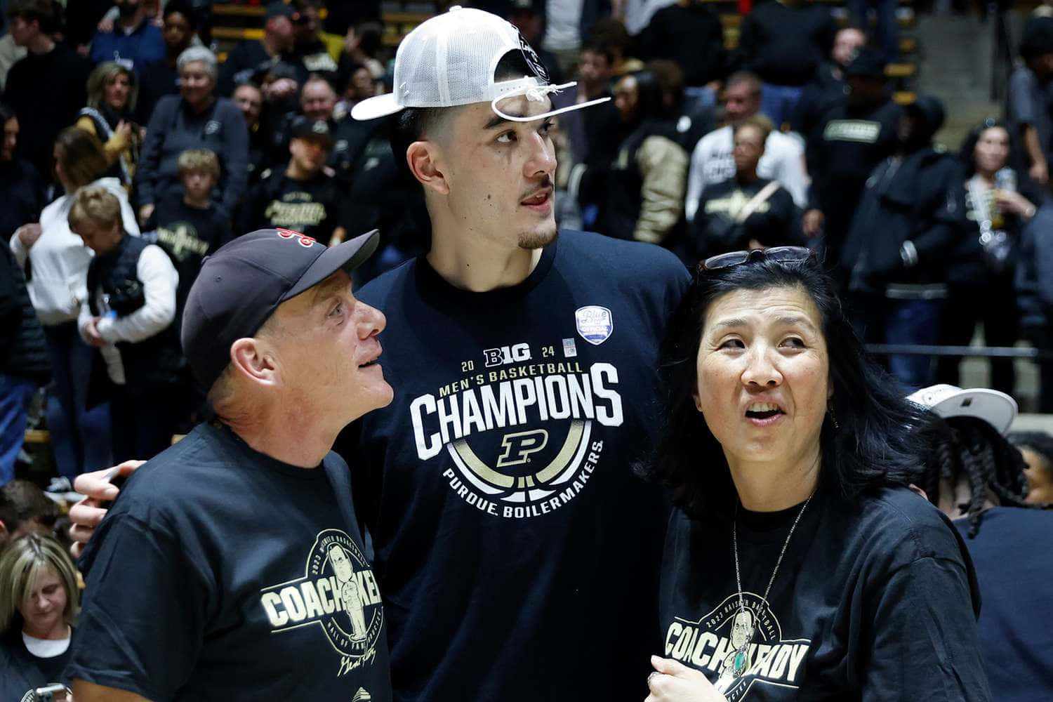  壁紙 — 一位身穿『Champions』球衣的勝利籃球運動員，站在微笑的教練和母親之間，身處擠滿觀眾的體育館。