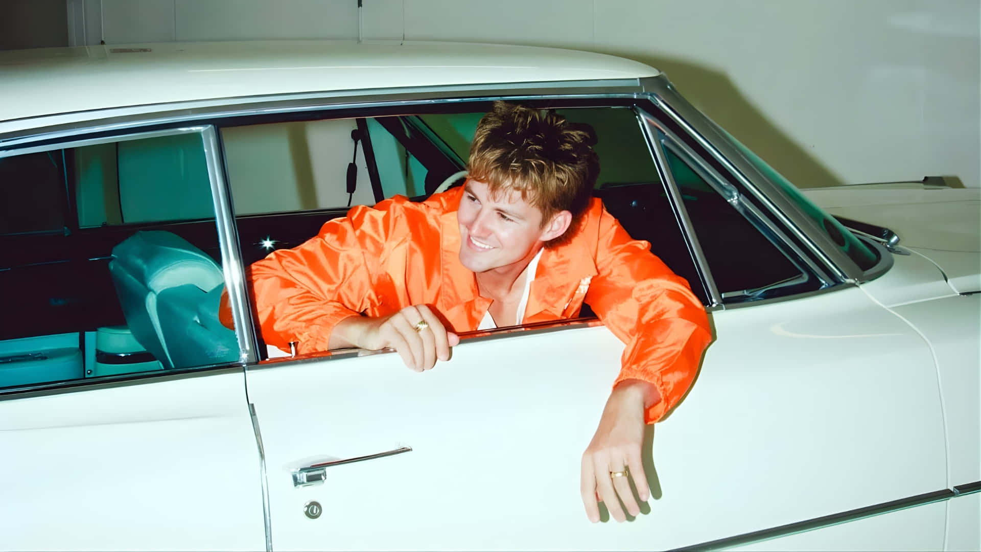  壁紙 — 一位身穿亮橙色外套的年輕男子，從白色經典汽車的車窗探出頭來，露出燦爛的笑容。