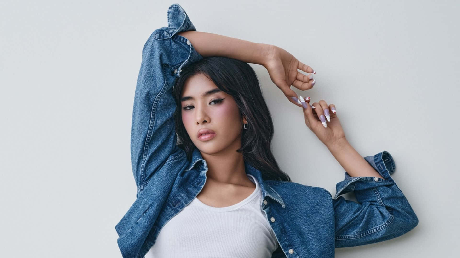  壁紙 — 一位深色頭髮、紫色指甲的年輕女性，身穿白色上衣外搭牛仔外套，在純白背景前自信地擺姿勢。