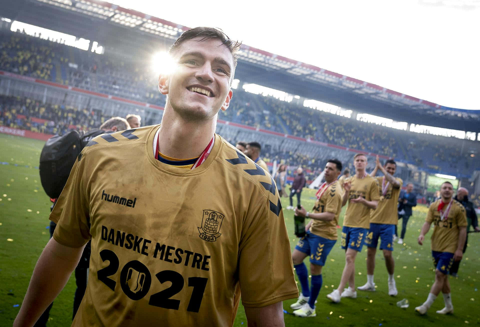  壁紙 — 一位勝利的足球運動員身穿金色 'Danske Mestre 2021' 球衣，在球場燈光下，隊友們在灑滿五彩紙屑的場地上慶祝，他笑容滿面。