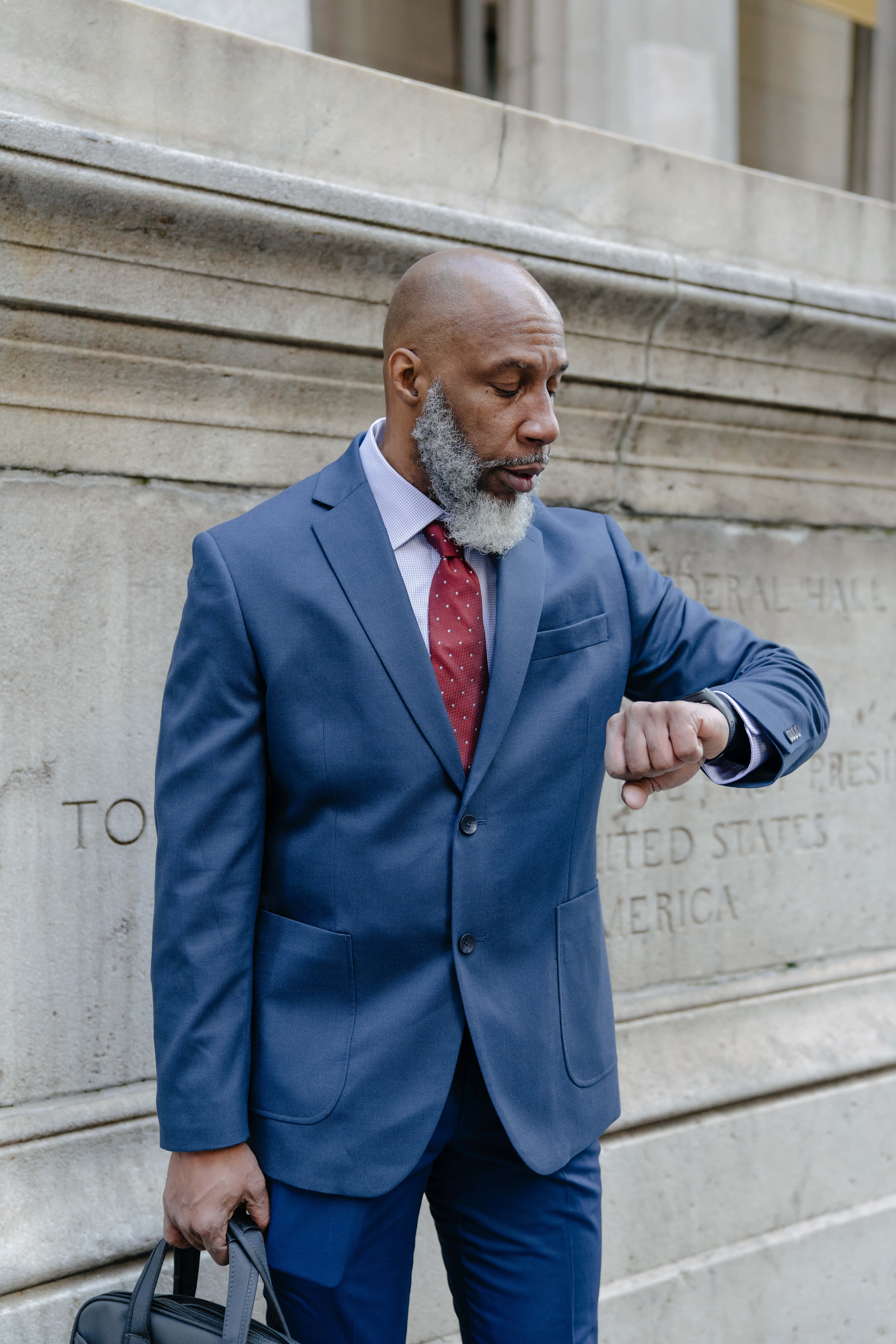 Navy Blue Blazer 桌布 壁紙 — 一位禿頭、灰色鬍鬚、身穿藍色西裝的男子，在石建築外牆前查看手錶。