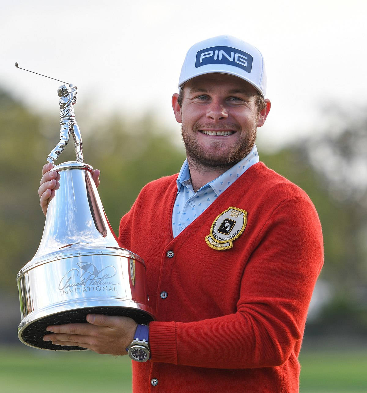Tyrrell Hatton 桌布 壁紙 — 一位微笑的高爾夫球手穿著紅色開襟衫，在模糊的綠色背景前，手持一個帶有高爾夫球手雕像的大型銀色獎盃。