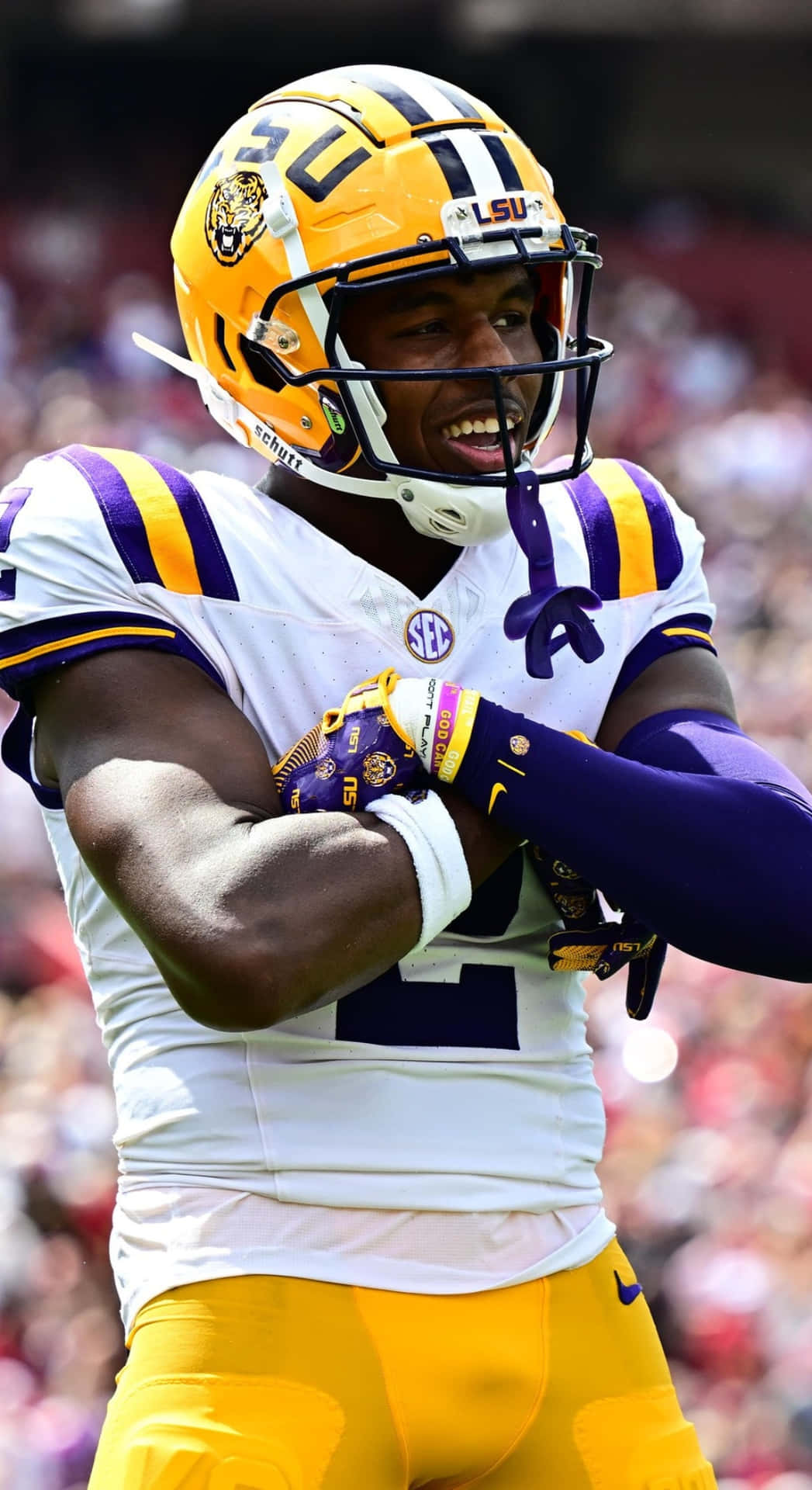  壁紙 — 一位微笑的路易斯安那州立大學（LSU）足球員，身穿黃紫色球衣，在陽光明媚的球場上交叉雙臂慶祝。