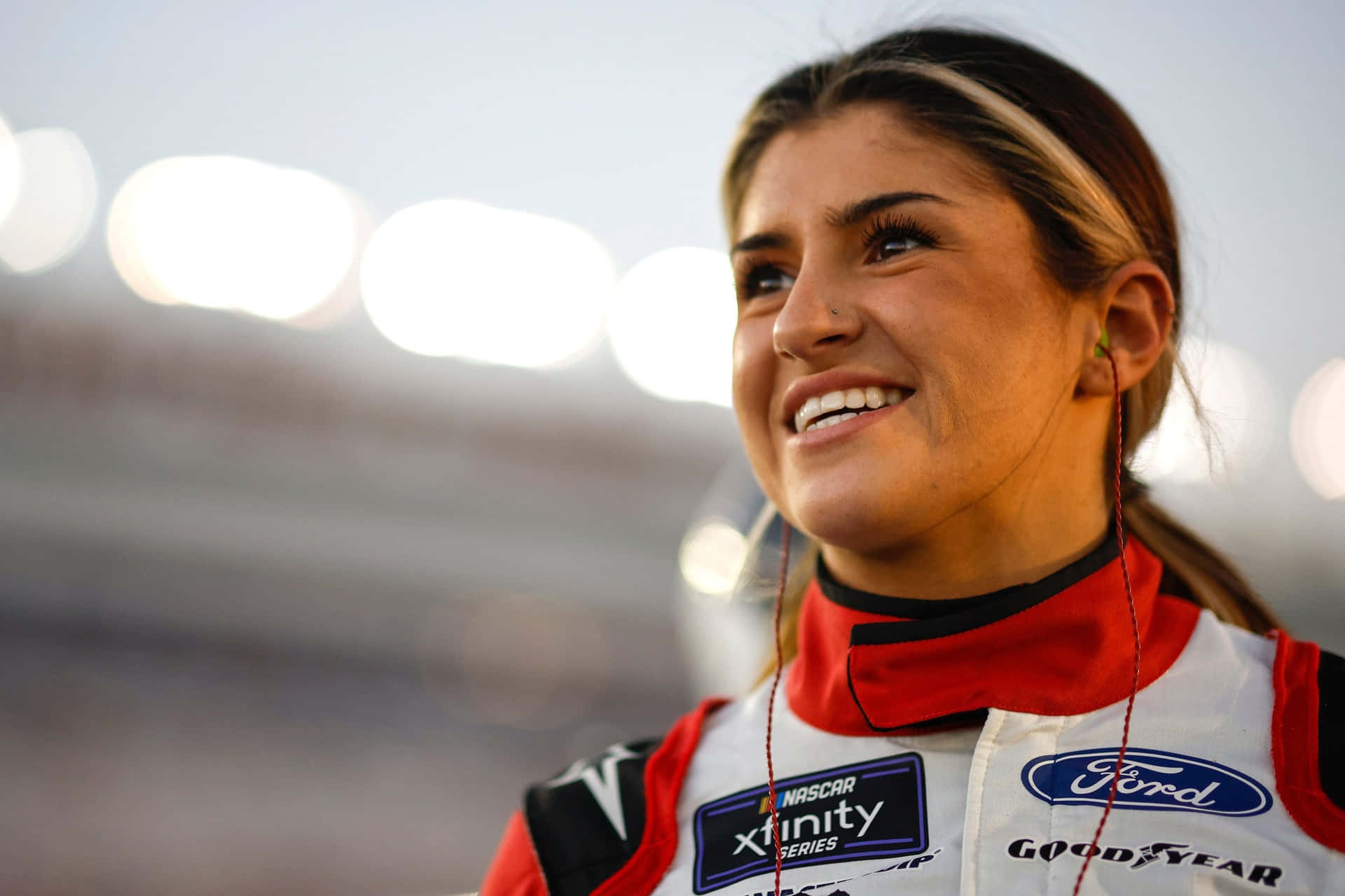  壁紙 — 一位微笑的女NASCAR車手穿著紅白黑賽車服，上有福特和固特異標誌，在模糊的體育場背景和明亮燈光下向上看。