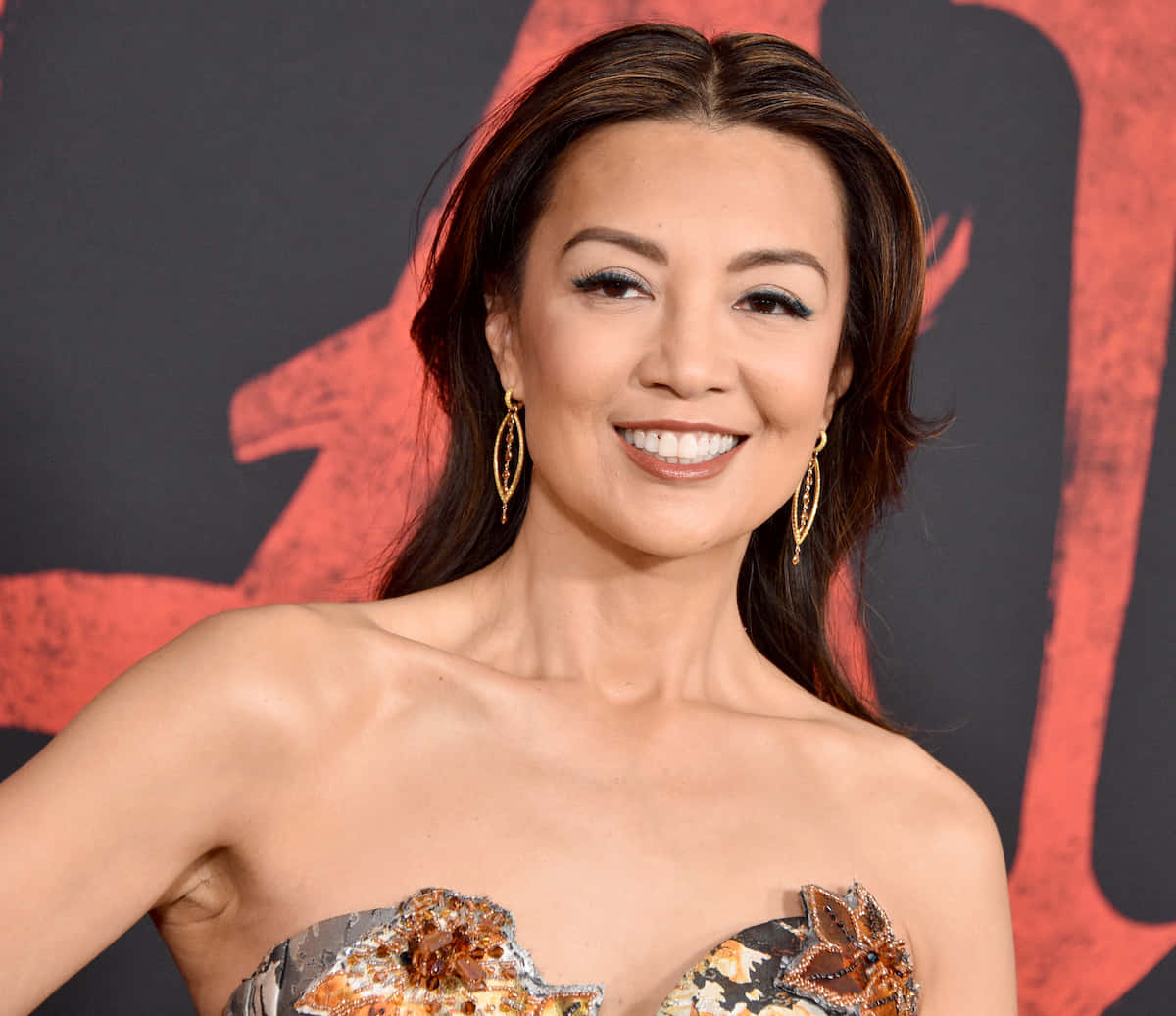 Ming Na Wen 桌布 壁紙 — 一位微笑的女性身穿鑲有寶石的露肩禮服，在鮮明的紅黑圖形背景前擺姿勢。
