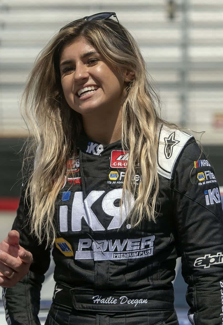  壁紙 — 一位微笑嘅女性NASCAR車手，身穿黑色賽車服，上面有贊助商標誌，自信咁企喺賽道上。