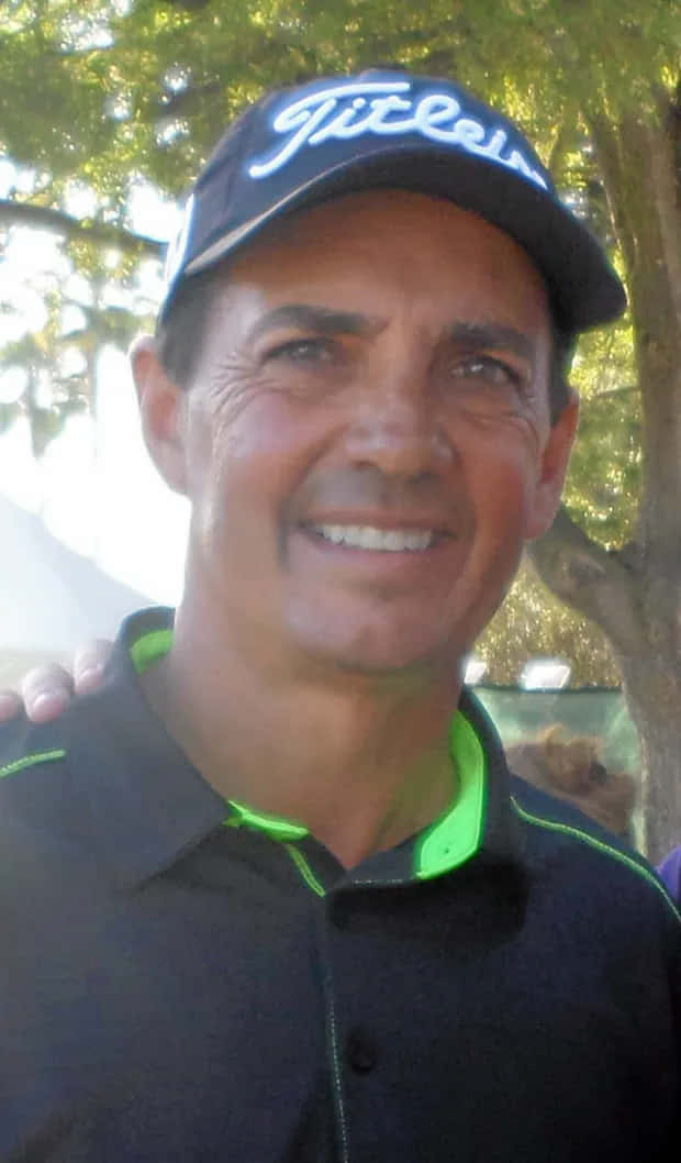  壁紙 — 一位微笑的男子戴著Titleist帽子和黑色Polo衫，帶有螢光綠色飾邊，站在戶外樹蔭下。