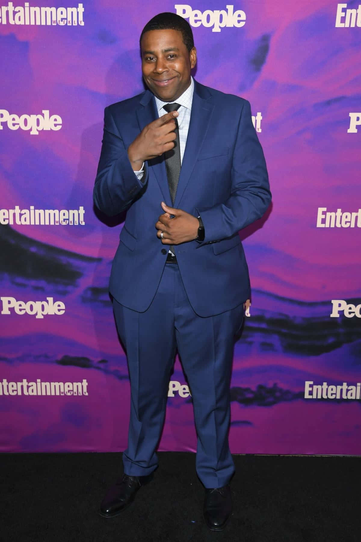 Purple Suit 桌布 壁紙 — 一位微笑的男子身穿俐落藍色西裝，在紫色「People Entertainment」品牌背景前自信擺姿勢。