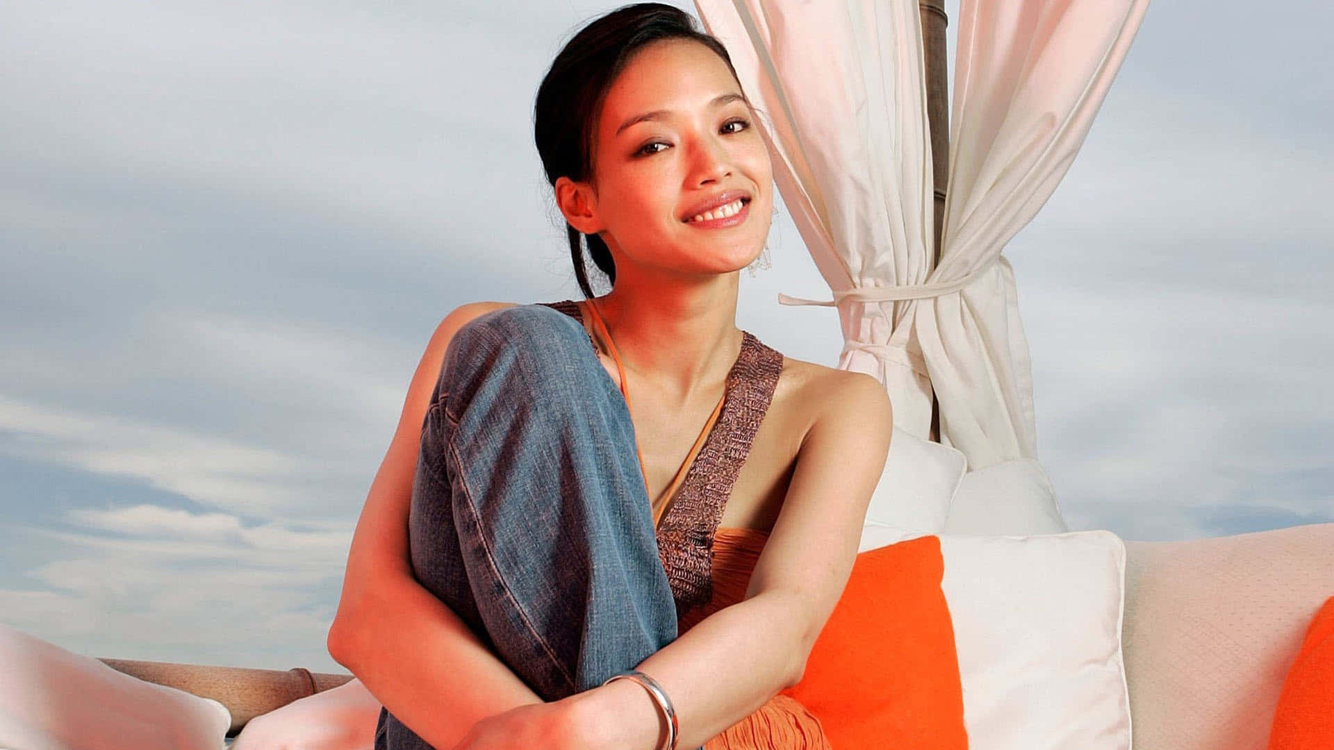 Shu Qi 桌布 壁紙 — 一位微笑的女性穿著休閒服裝，在柔和天空下放鬆地坐在有軟墊的戶外躺椅上。