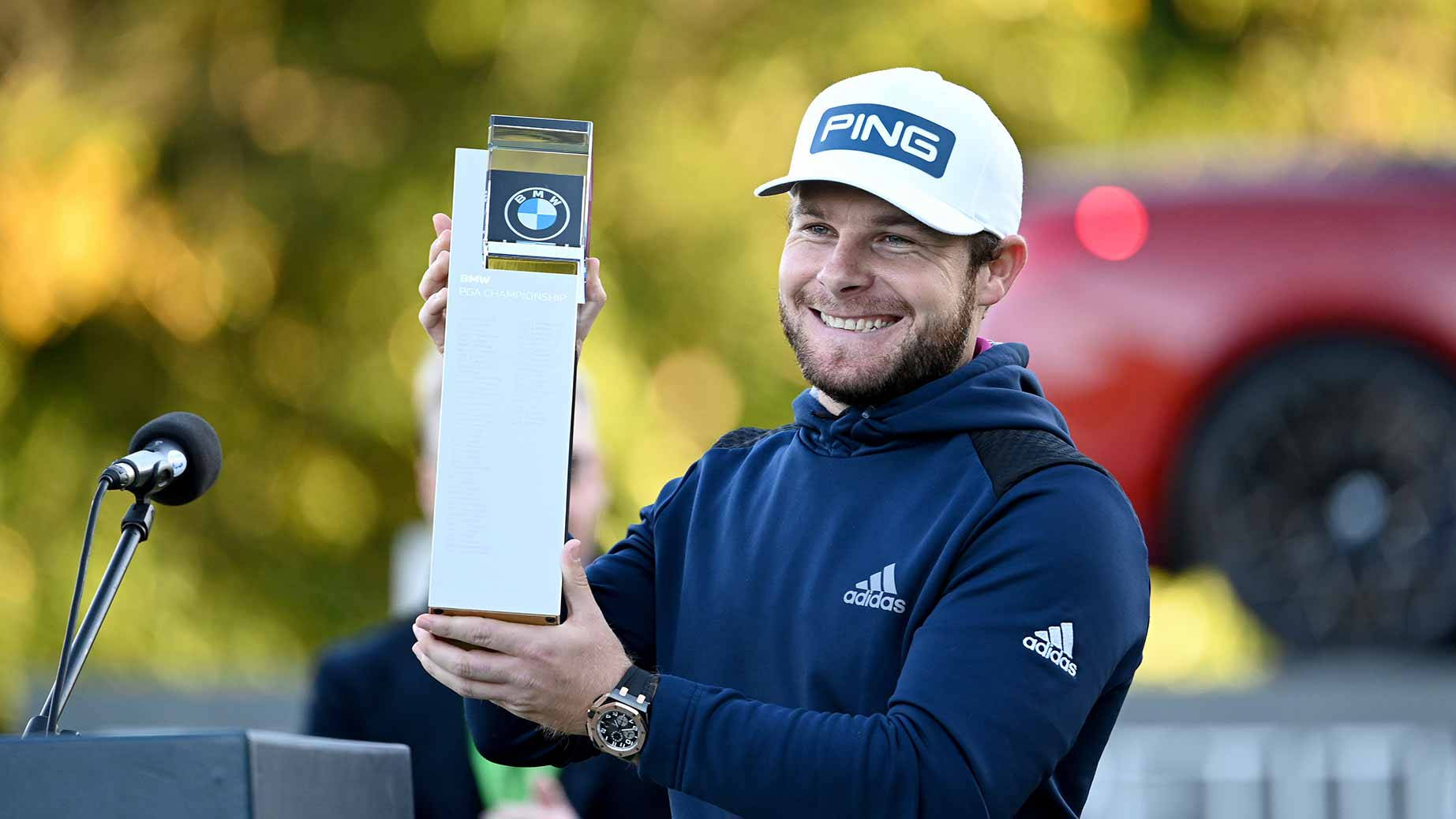Tyrrell Hatton 桌布 壁紙 — 一位微笑的高爾夫球手穿著藍色連帽衫和白色帽子，在頒獎台上自豪地舉起BMW獎盃，背景是模糊的紅色汽車和綠色樹葉。