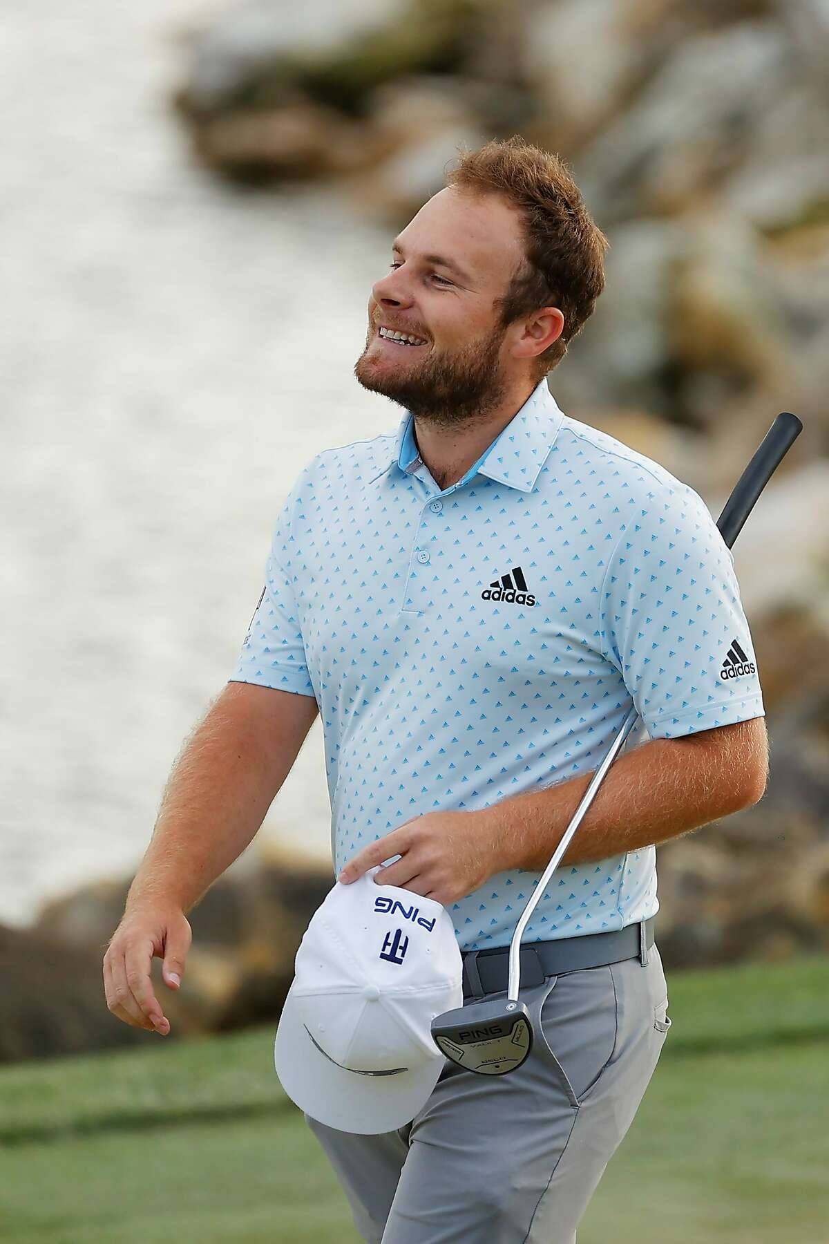 Tyrrell Hatton 桌布 壁紙 — 一位微笑的高爾夫球手穿著淺藍色圖案襯衫，在草地上行走，手持白色帽子和球桿。