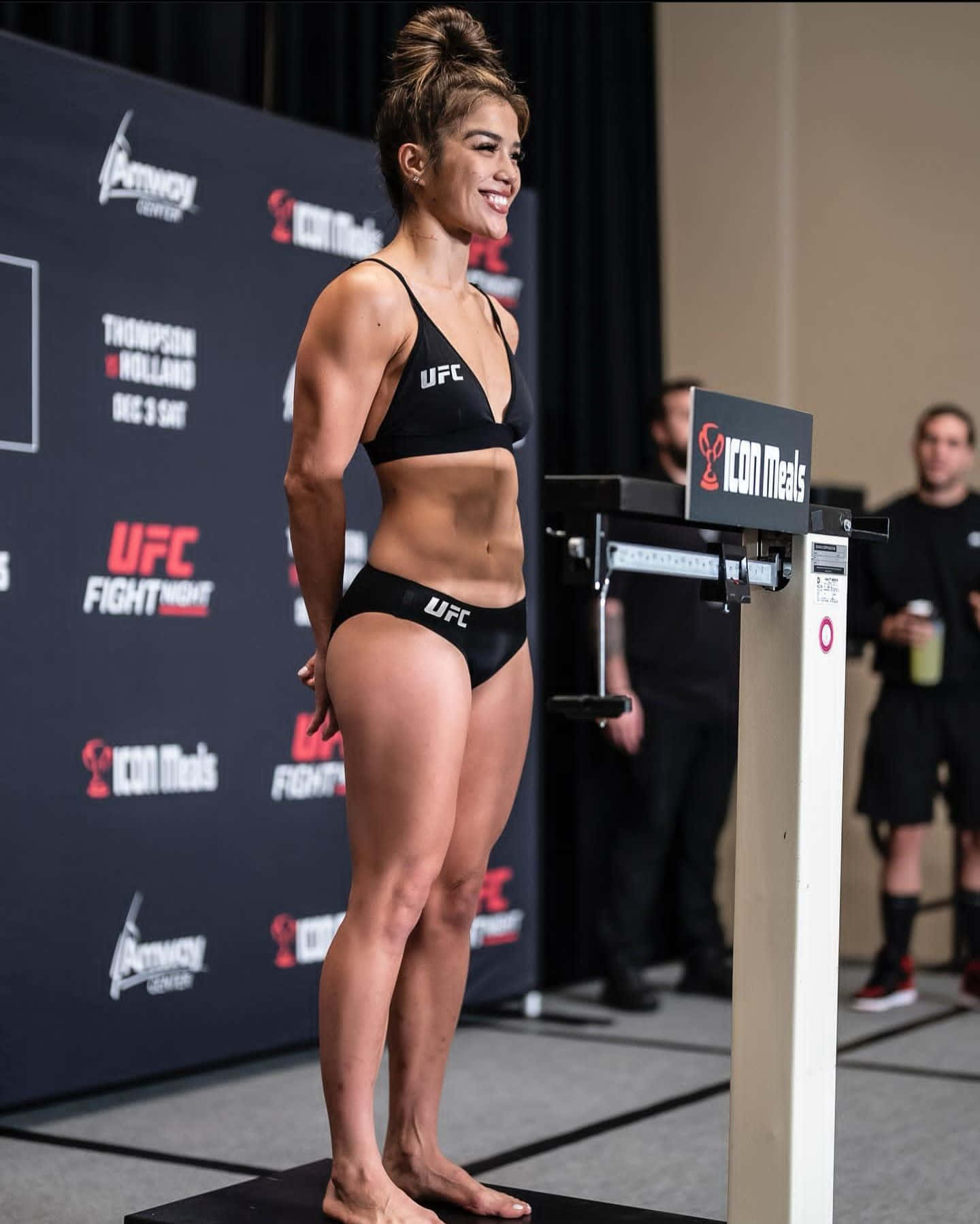  壁紙 — 一位微笑的女性UFC拳手身穿黑色運動裝備，自信地站在有品牌背景的稱重活動的體重秤上。