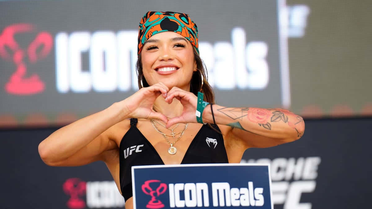  壁紙 — 一位微笑的女拳手身穿黑色UFC上衣，在'ICON Meals'品牌背景的稱重活動中用手比出心形。