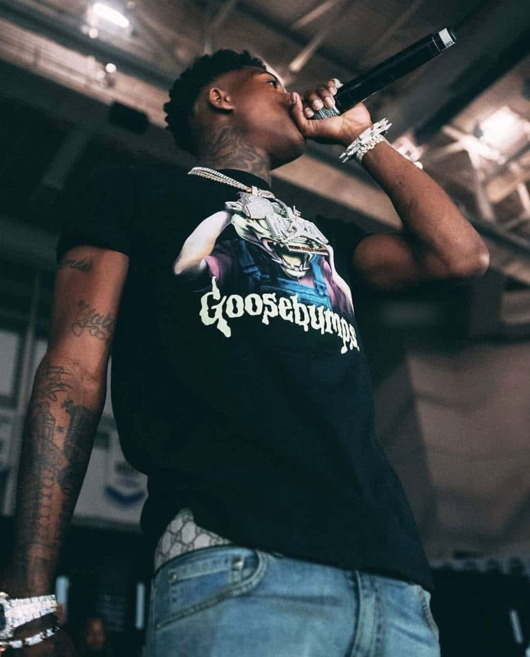 NBA Youngboy 在聚光燈下盡情享受 壁紙 — 一位紋身說唱歌手穿著 Goosebumps T恤，在昏暗的演唱會燈光下熱情地對著麥克風表演。