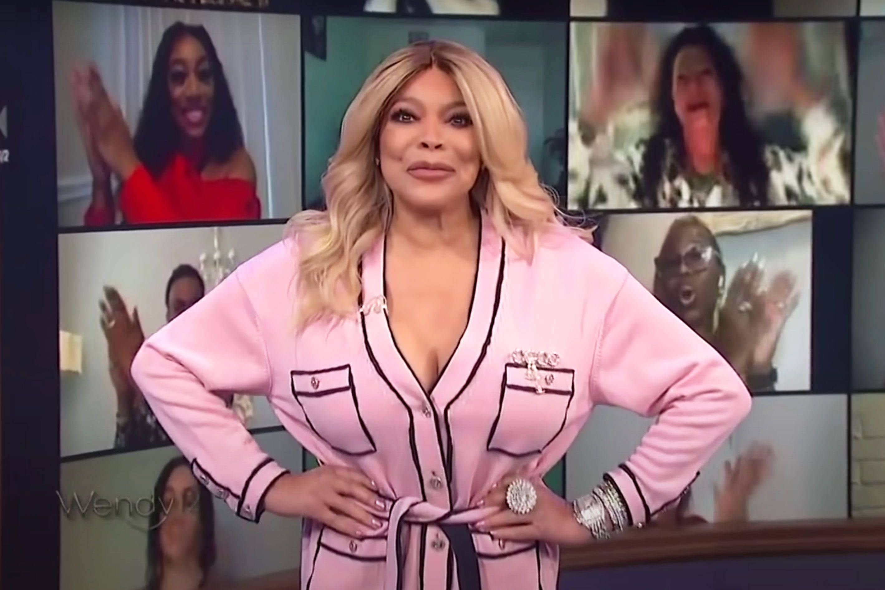 Wendy Williams Before A Screen 壁紙 — Wendy Williams 自信地穿著帶黑色飾邊的粉紅色長袍，身後的多個視頻屏幕顯示著歡呼的人群。