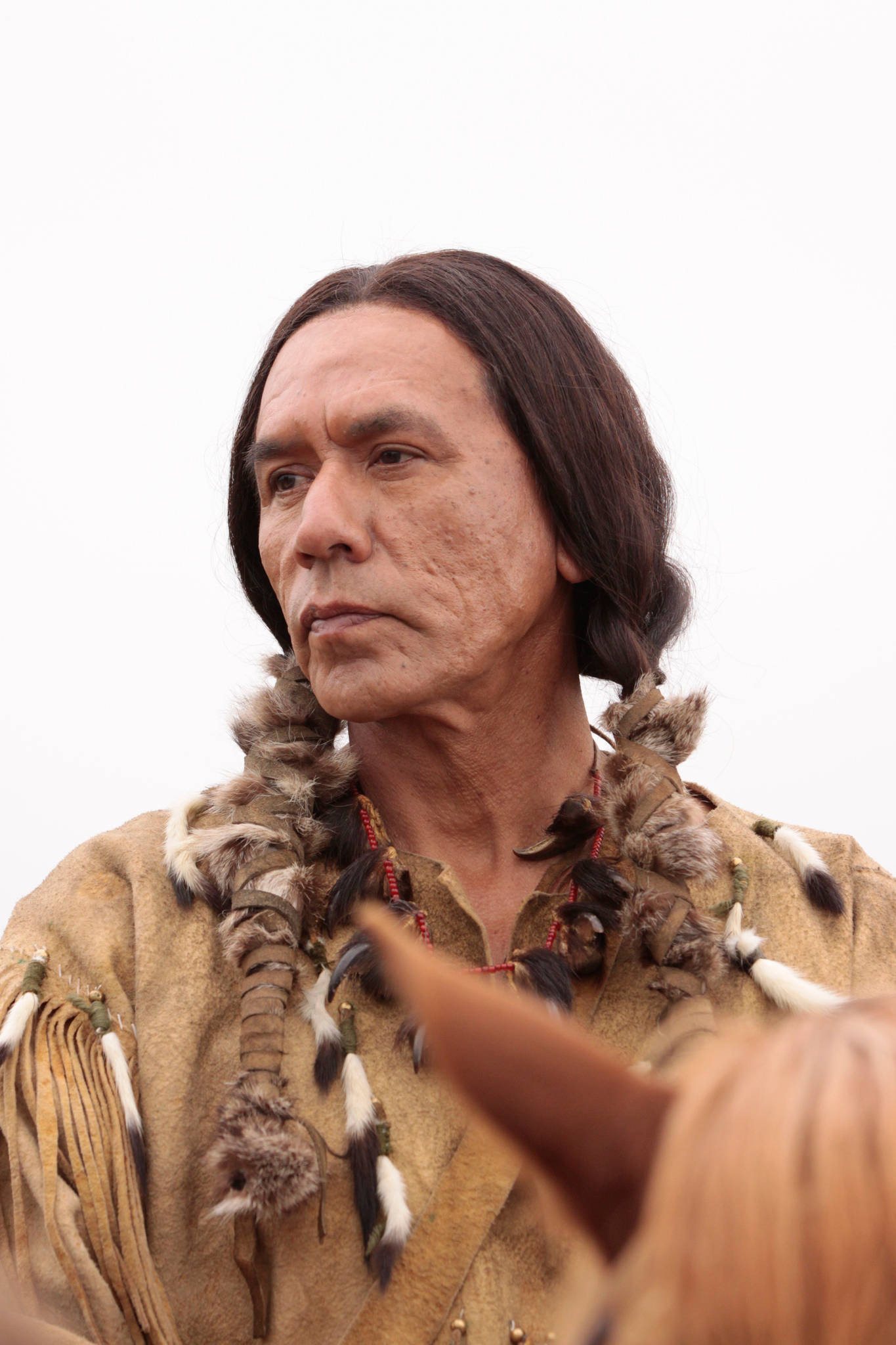 Wes Studi 在電影《Hell on Wheels》 壁紙 — 一位身穿傳統羽毛服飾的美洲原住民男子在蒼白的天空下，若有所思地凝視著馬匹旁邊。