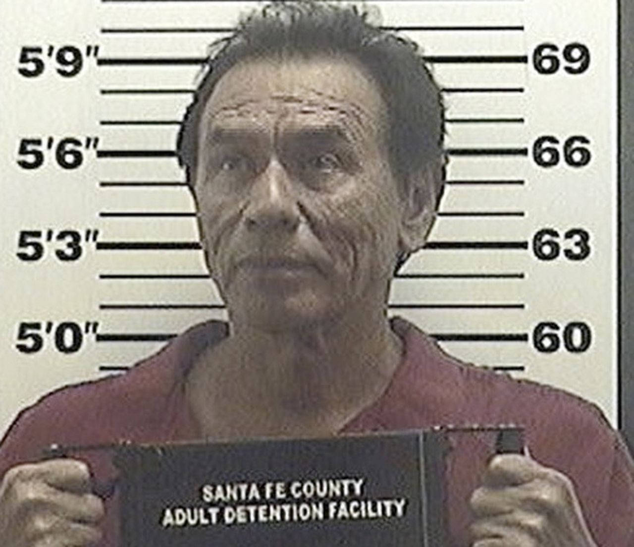 Wes Studi 被逮捕 壁紙 — 一位身穿紅色連身衣的長者站在身高尺前，手持寫有聖塔菲縣成人拘留所的標誌。